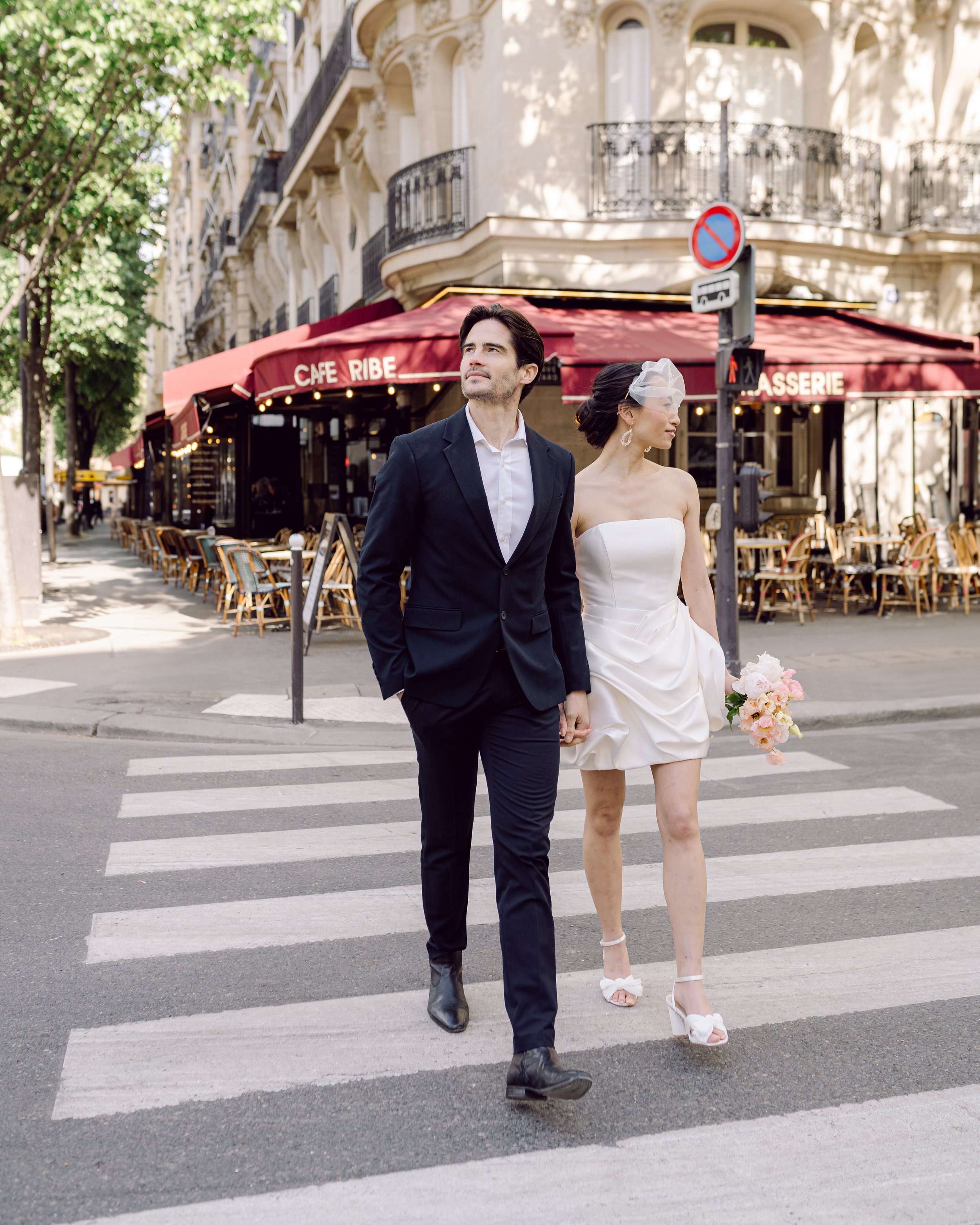 Un couple marchant sur un passage piéton à Paris. La femme porte une robe de mariée courte et des talons hauts, tenant un bouquet de fleurs. L'homme porte un costume noir. Un café avec terrasse et des bâtiments haussmanniens sont en arrière-plan.