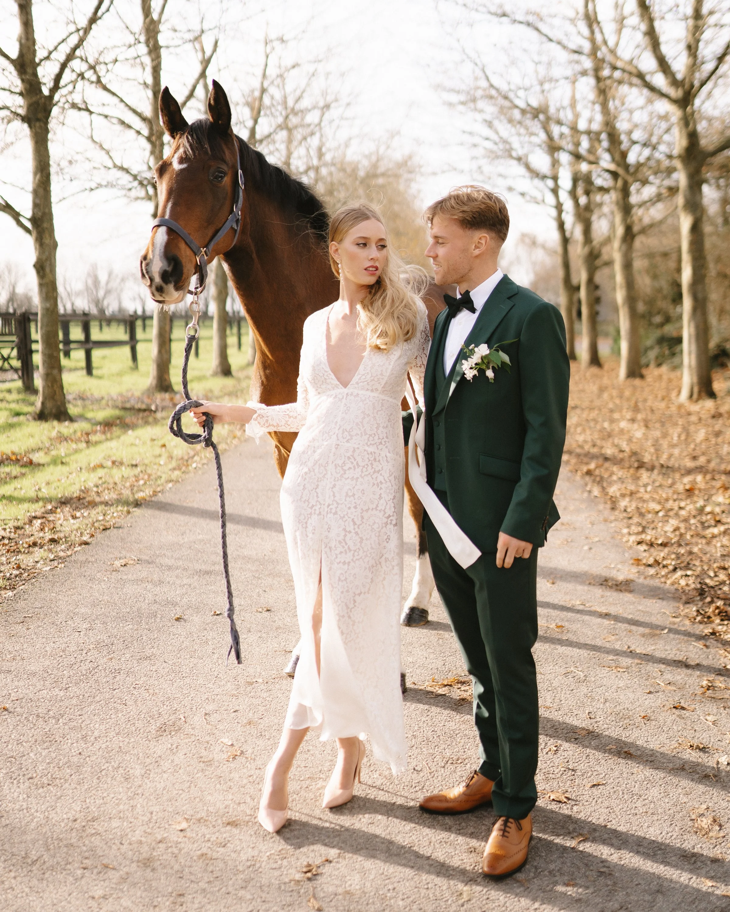 Un couple en tenue de mariage, la femme en robe longue blanche et l'homme en costume vert foncé avec nœud papillon, se tiennent sur un chemin bordé d'arbres, avec un cheval derrière eux.