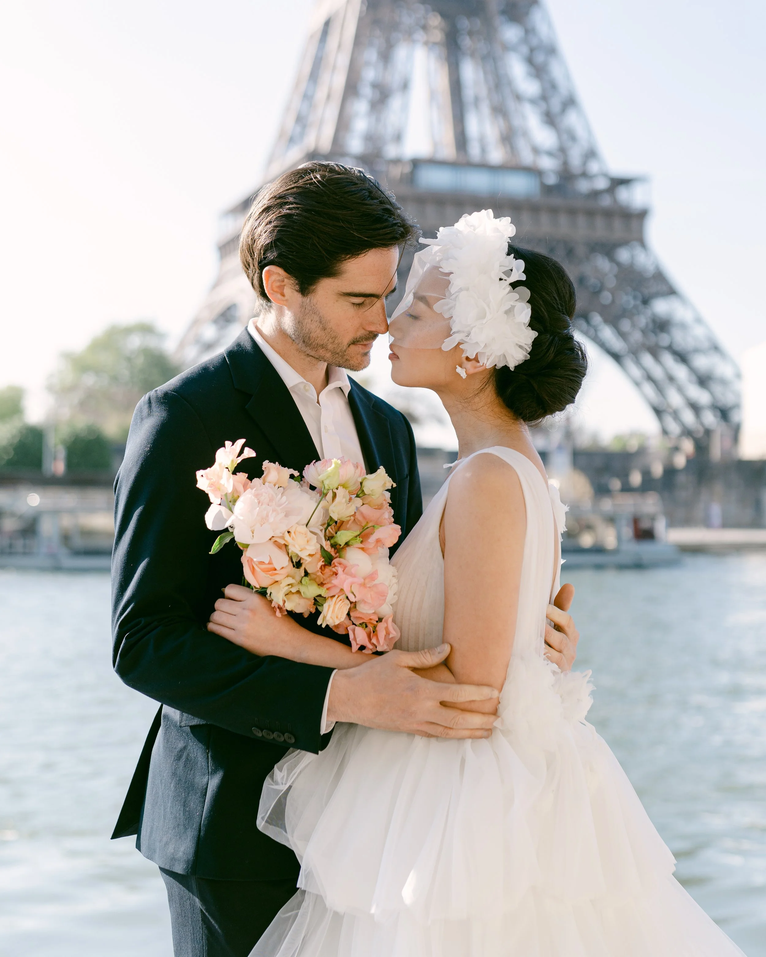 Un couple de mariés à Paris, devant la tour Eiffel, se regardant tendrement, la mariée portant une robe blanche avec un voile et un bouquet de fleurs, le marié en costume noir, près de la Seine.