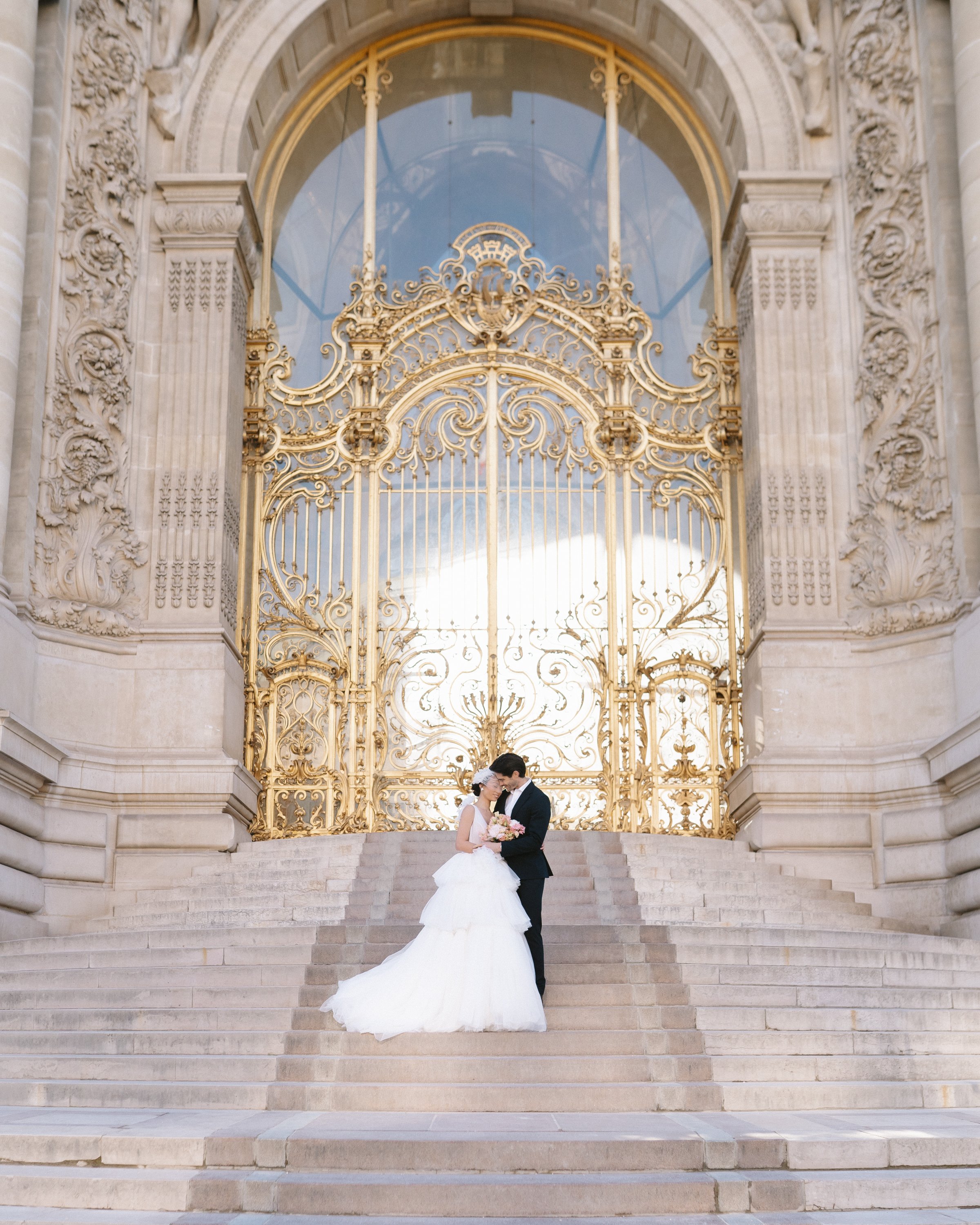 Un couple de mariés se tenant par la taille en haut des marches d'une entrée ornée d'une grille en fer doré, avec un bâtiment en pierre en arrière-plan.