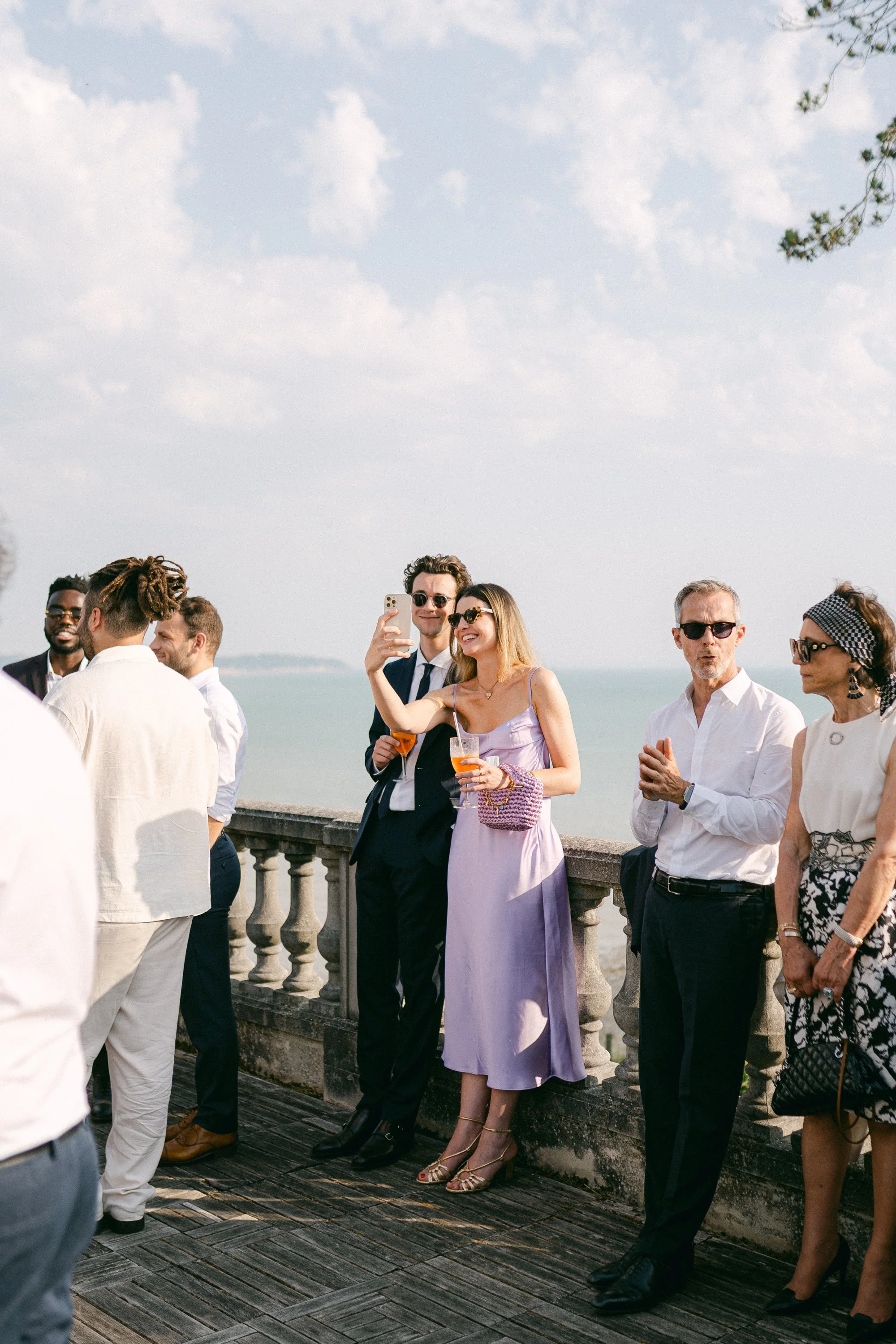 Groupe de personnes en tenue élégante, devant une mer ou un grand lac, prenant des photos et discutant lors d'un événement en plein air par une journée ensoleillée.