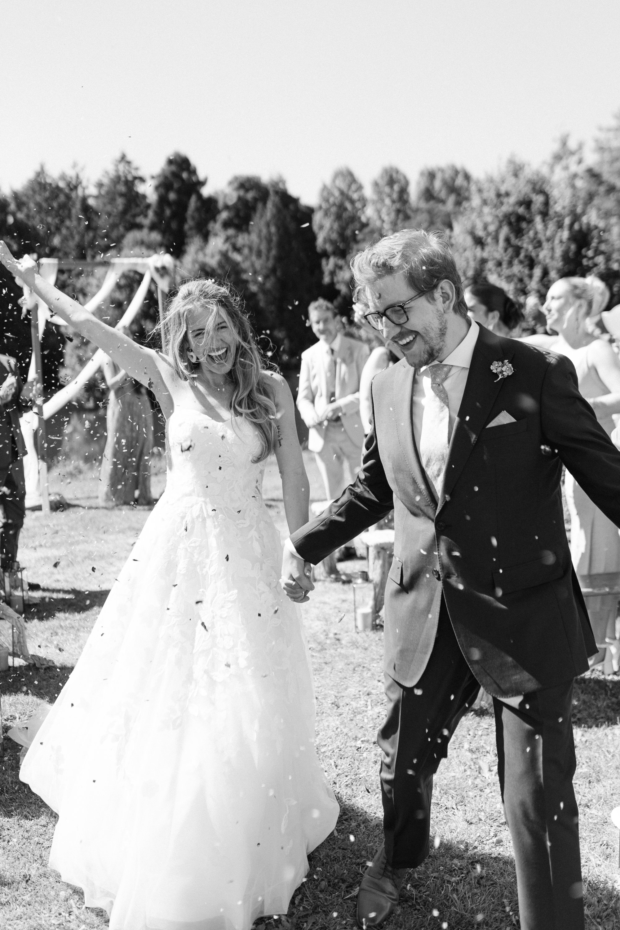 Un couple de mariés heureux lors de leur mariage en plein air, entourés de invités, avec des confettis, en noir et blanc.