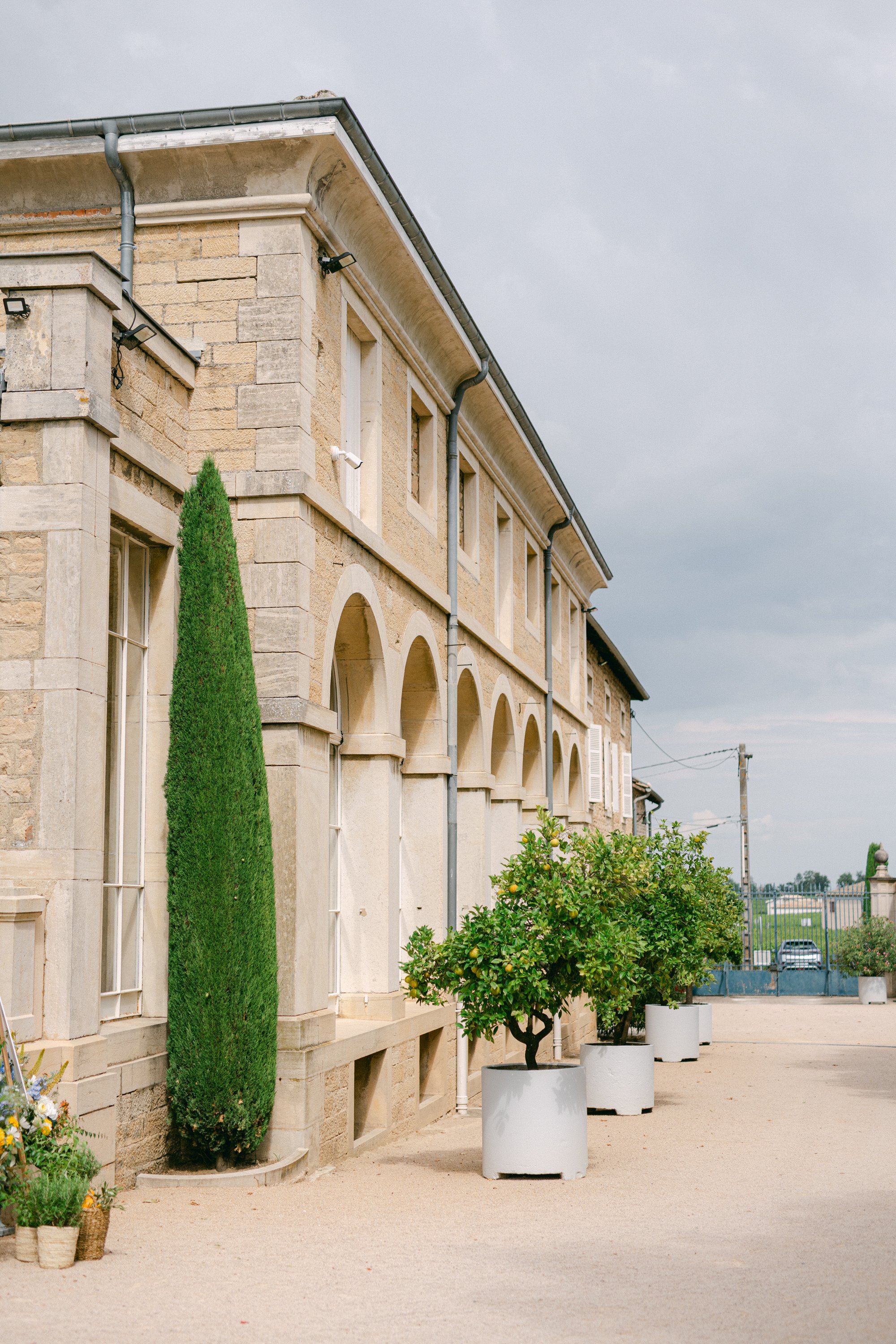 Facade en pierre d'un bâtiment avec fenêtres, plantes en pots alignés devant, ciel nuageux.
