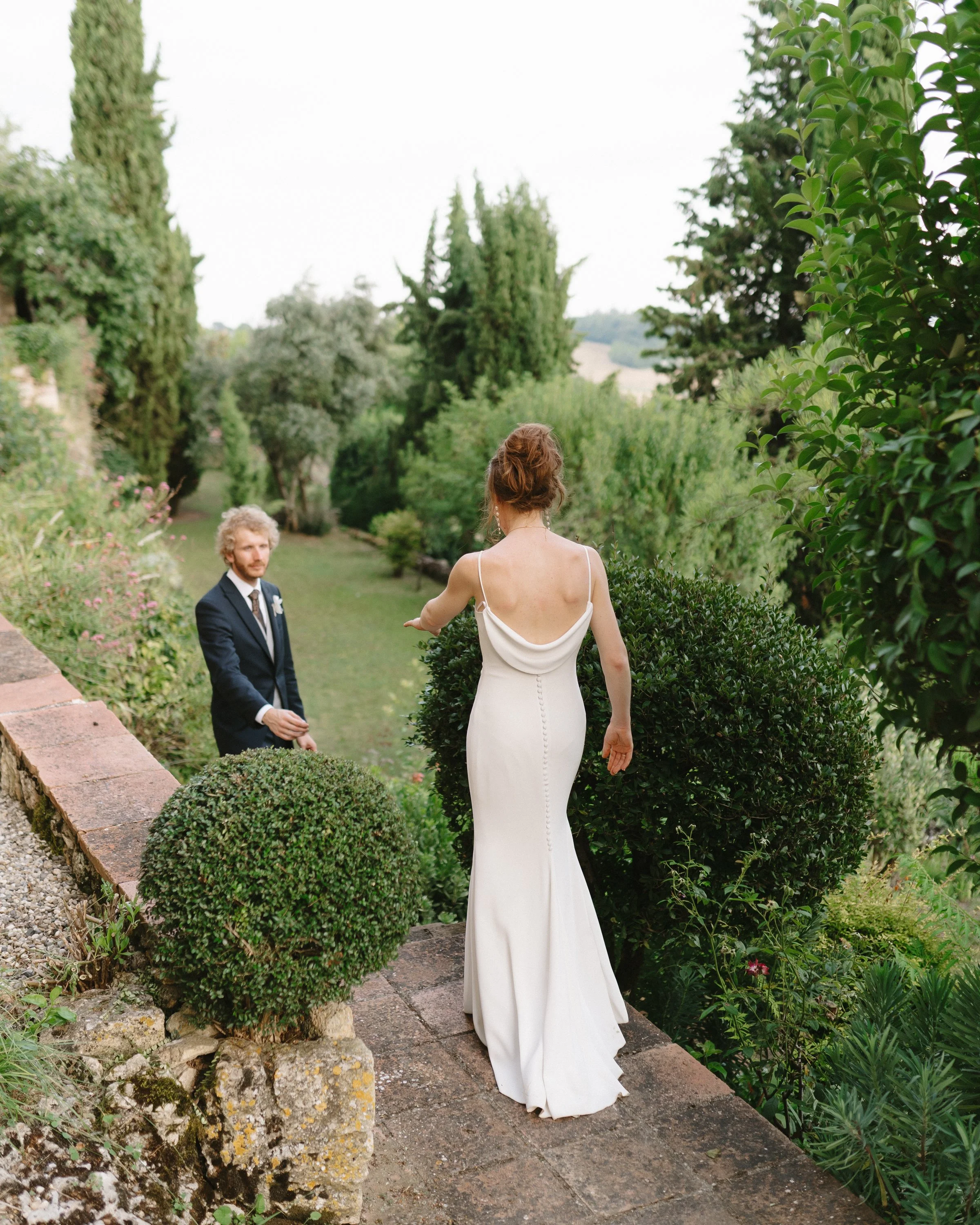 Une femme en robe blanche se tient sur un trottoir pavé dans un jardin, regardant un homme en costume noir qui se trouve plus bas dans le jardin, entouré de feuillage et d'arbres.