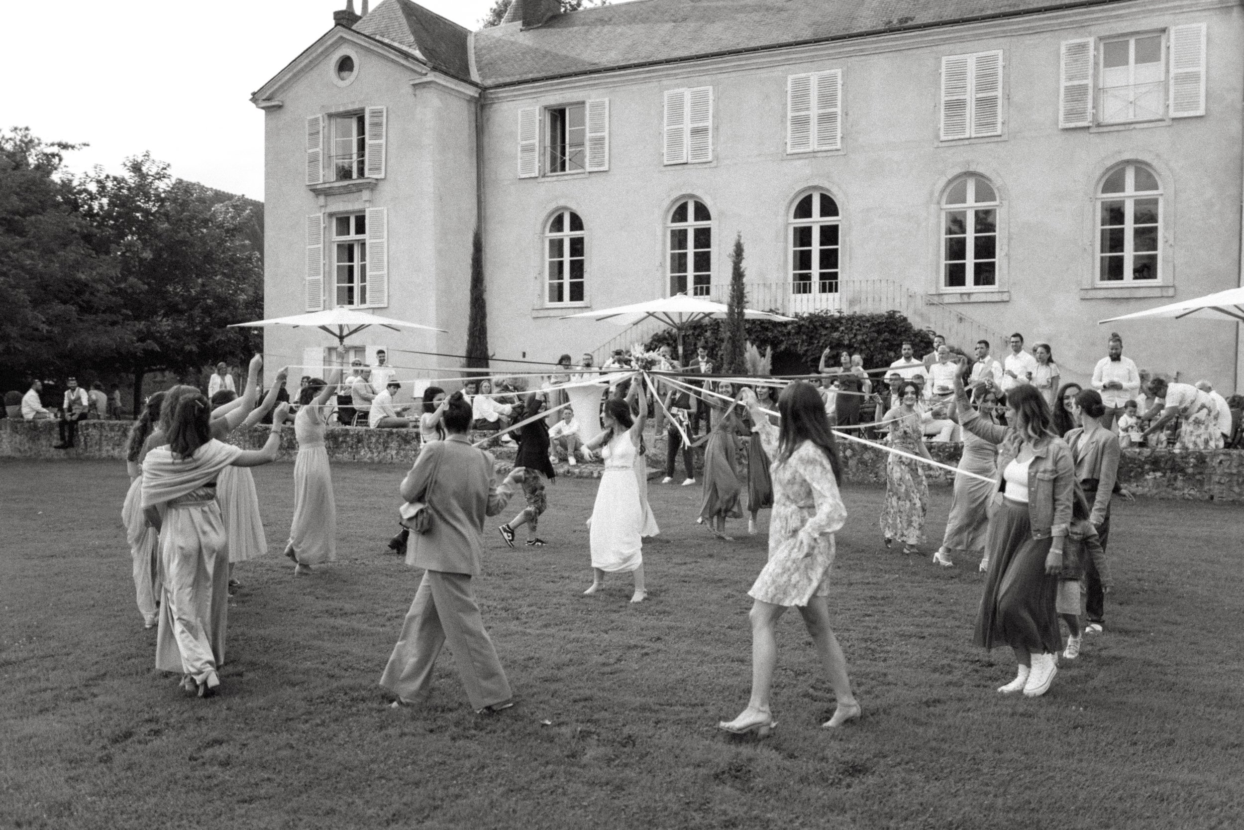 Groupe de personnes dansant en cercle avec des foulards sur une pelouse devant une grande maison ancienne avec parasols.
