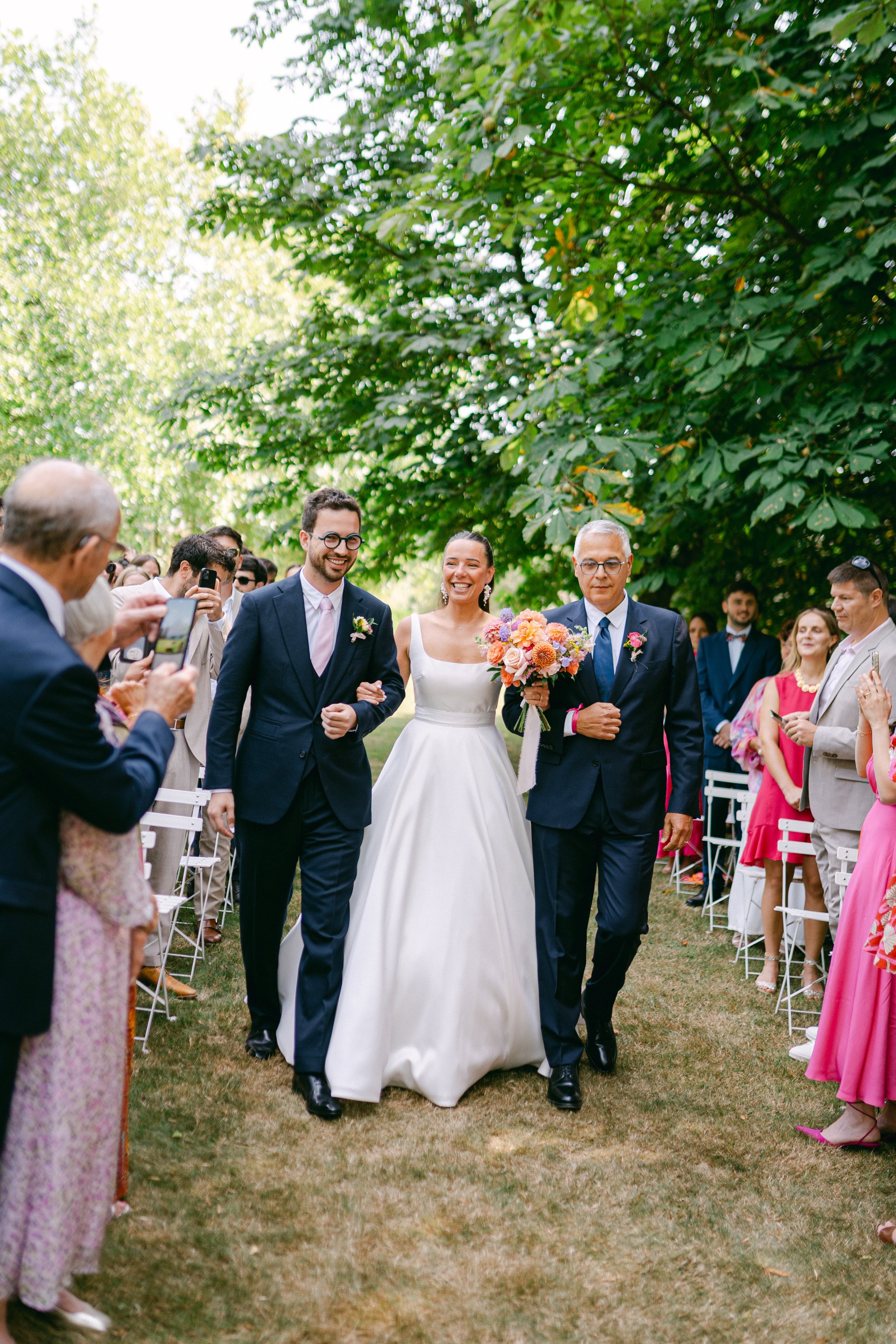 Un couple de mariés, la mariée avec une robe blanche, marche dans un jardin entouré de personnes en costume et en robes colorées, souriants, lors d'une cérémonie de mariage.