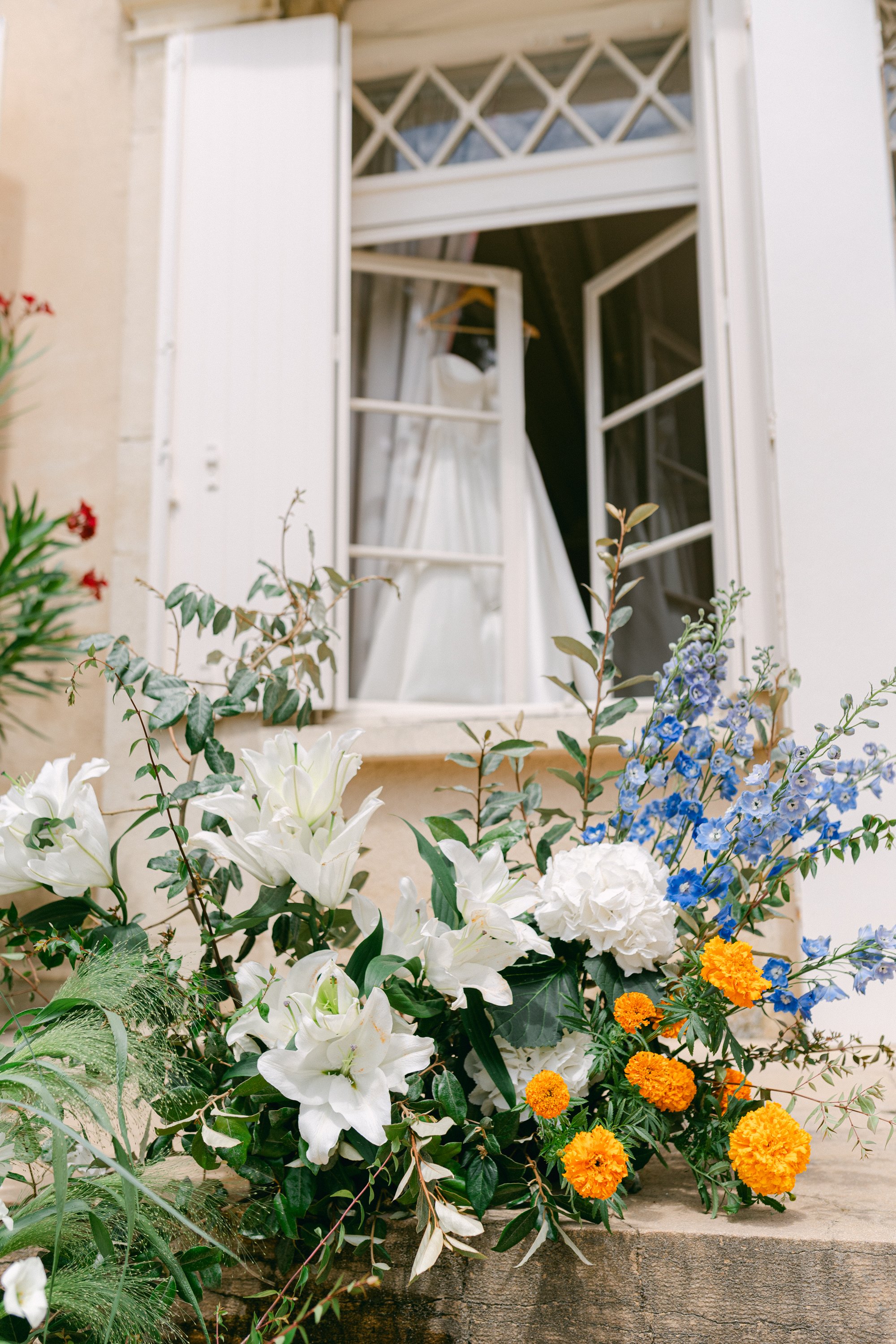 Fleurs colorées devant une fenêtre ouverte avec rideau blanc, située sur un mur beige.