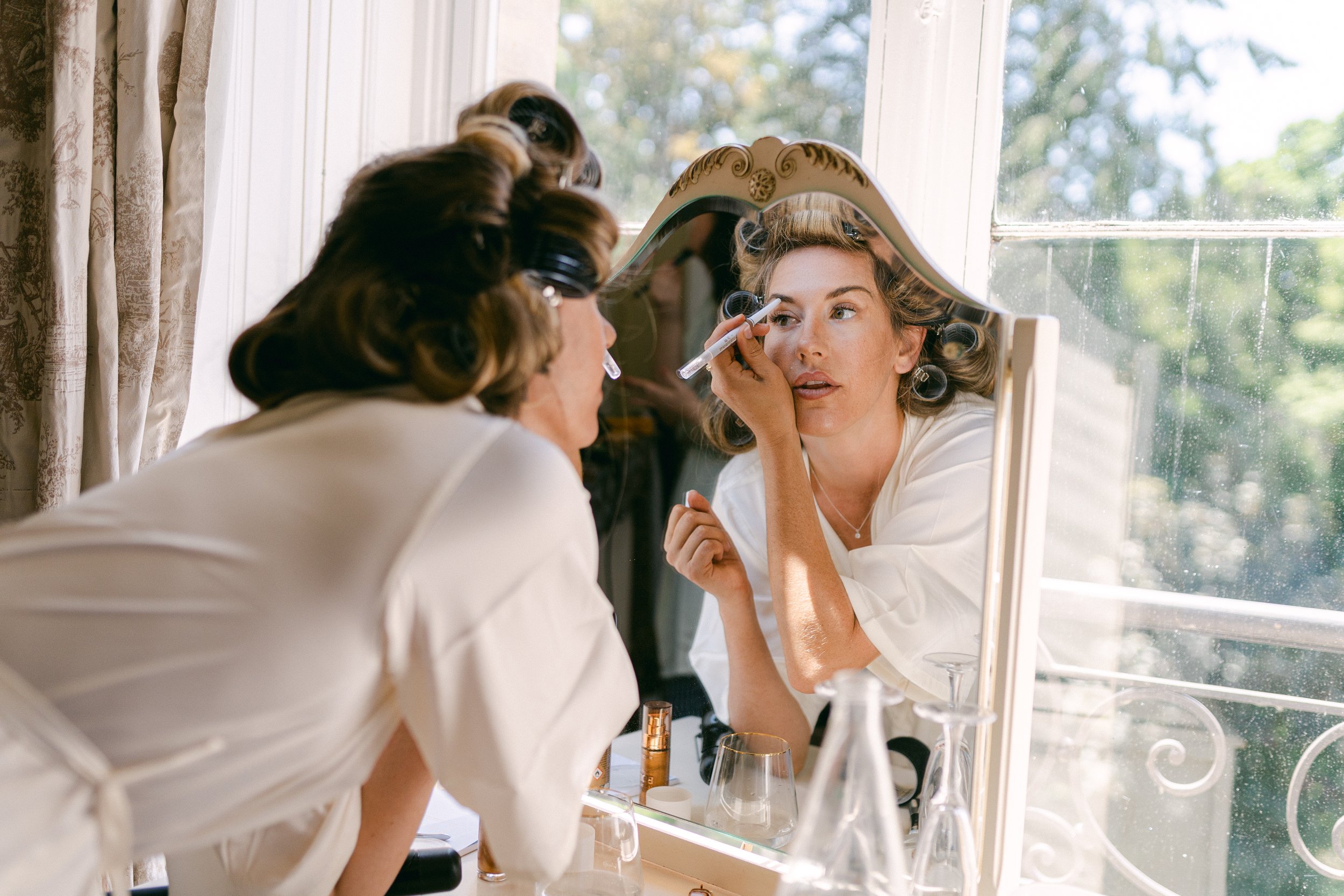 Une femme se maquille devant un miroir en s'appliquant du maquillage autour des yeux, avec une fenêtre lumineuse en arrière-plan.