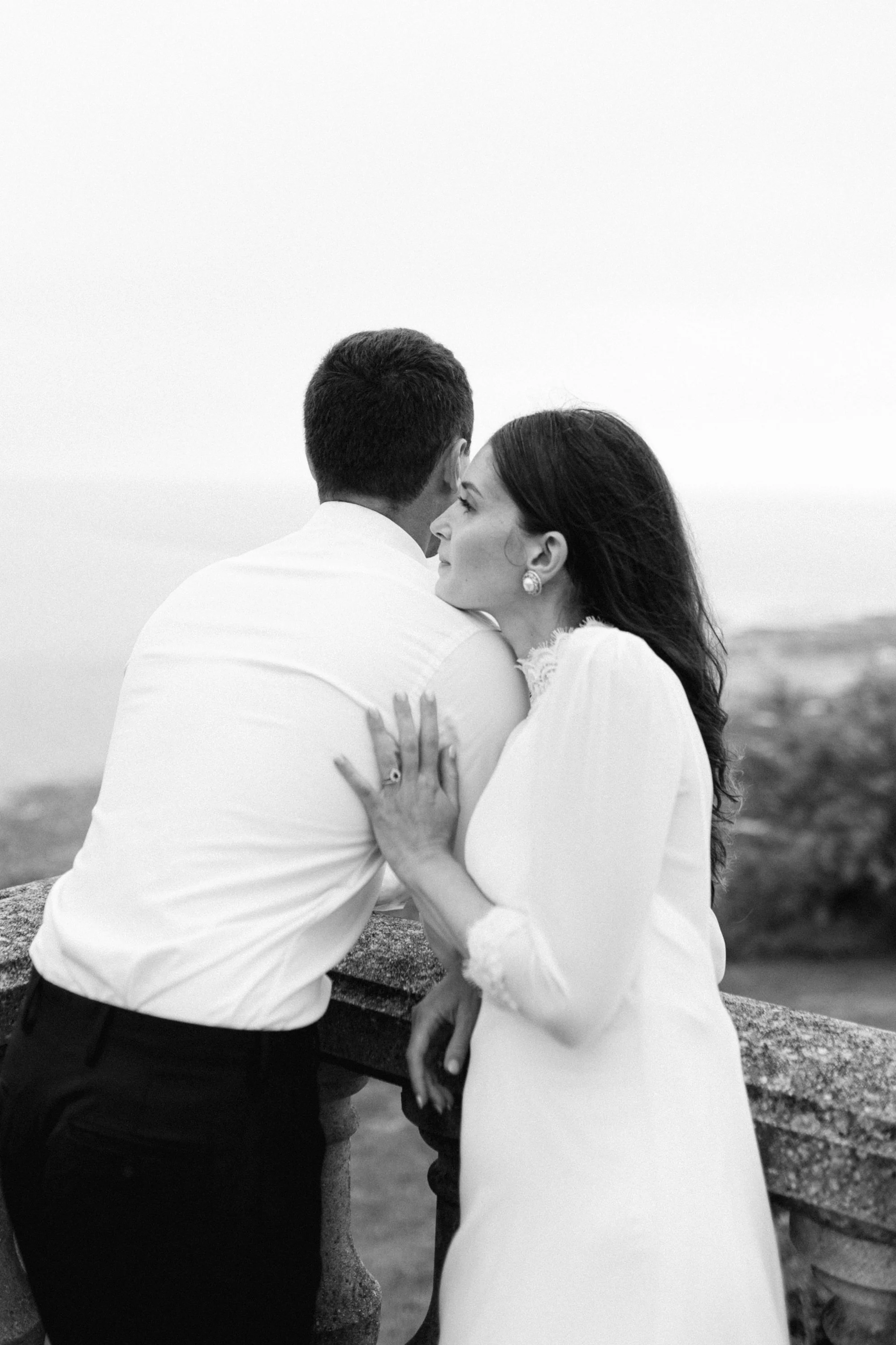 Un couple en noir et blanc, le homme vu de dos et la femme de profil, se tenant près d'une balustrade en pierre, avec un arrière-plan flou d'une vue panoramique.