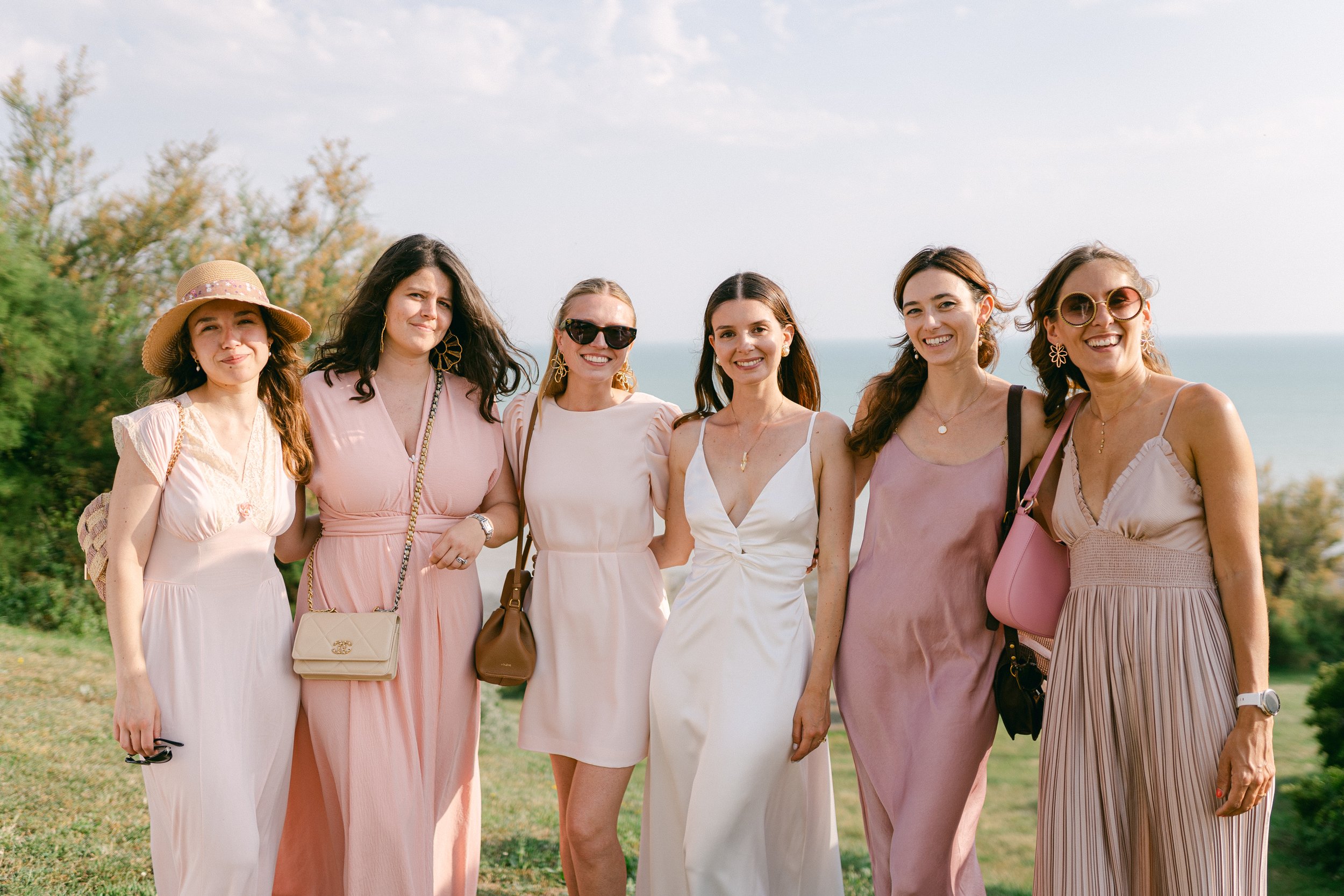 Groupe de six femmes souriantes posant ensemble en extérieur avec vue sur la mer, portant des vêtements d'été et des accessoires colorés.