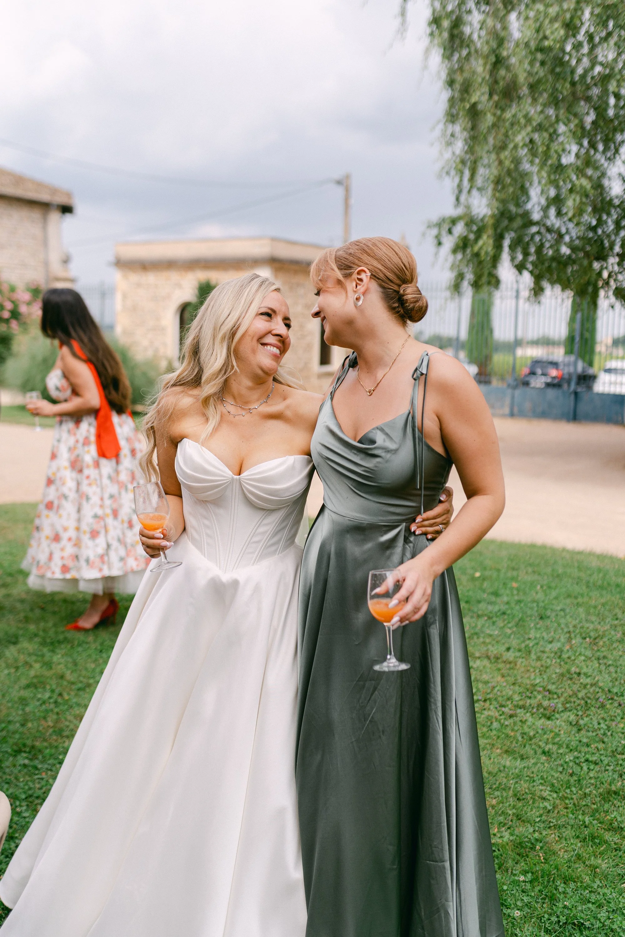Deux femmes en robes de soirée s'embrassent et sourient lors d'une fête en plein air, tenant des verres avec une boisson orange.