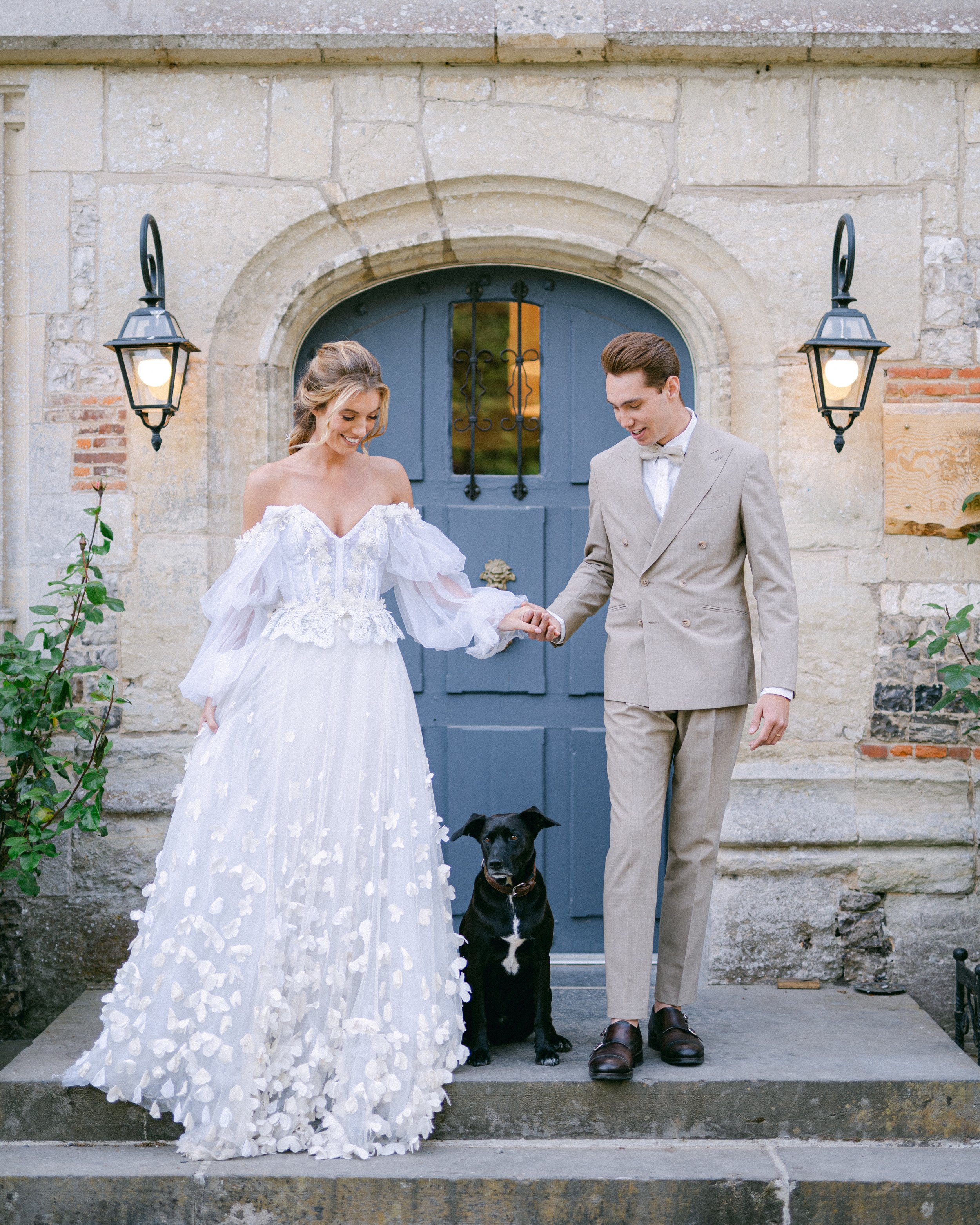 Un couple de mariés, une femme en robe de mariée blanche et un homme en costume beige, se tenant par la main devant une porte en bois bleue avec un chien assis entre eux, à l'extérieur d'un bâtiment en pierre.