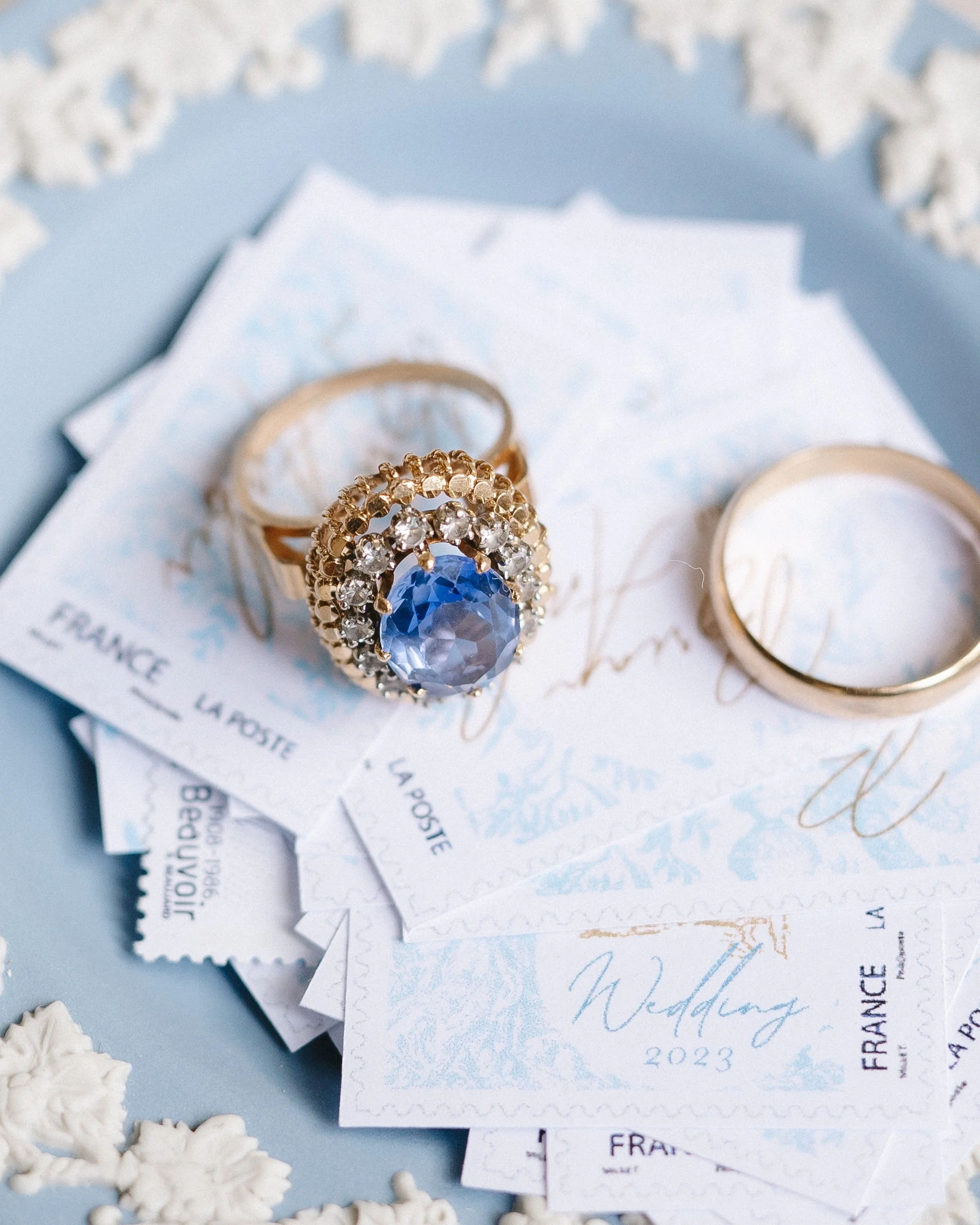Anneau en or avec une pierre précieuse bleue entourée de petits diamants, posé sur des papiers avec inscriptions en français, étiquettes de mariage. Ring en or et papier avec motifs floraux blancs en arrière-plan.