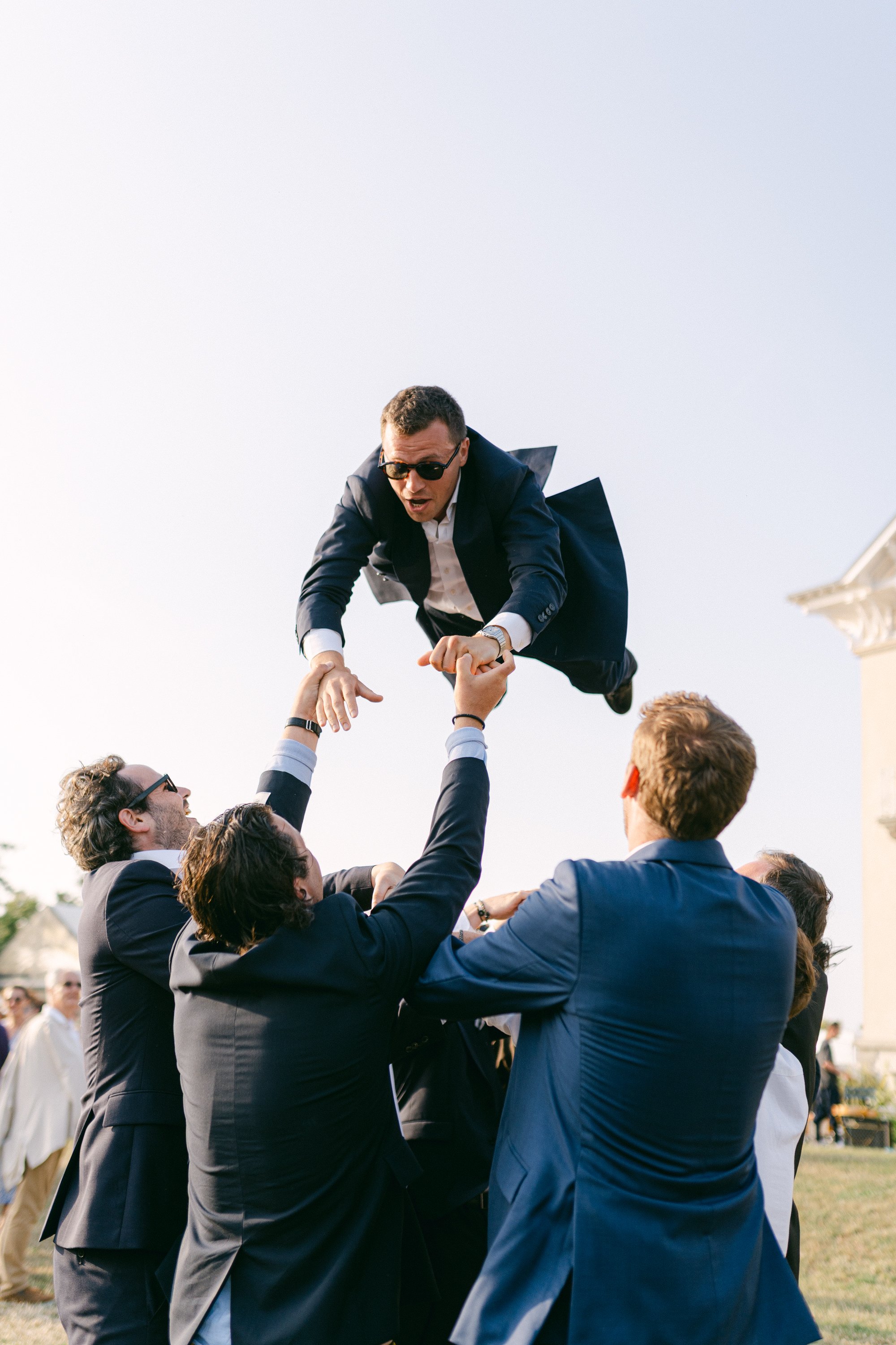Groupe de personnes en costume aidant un homme à le soulever dans les airs, il porte des lunettes de soleil et un costume bleu Marine, lors d'un événement en plein air.