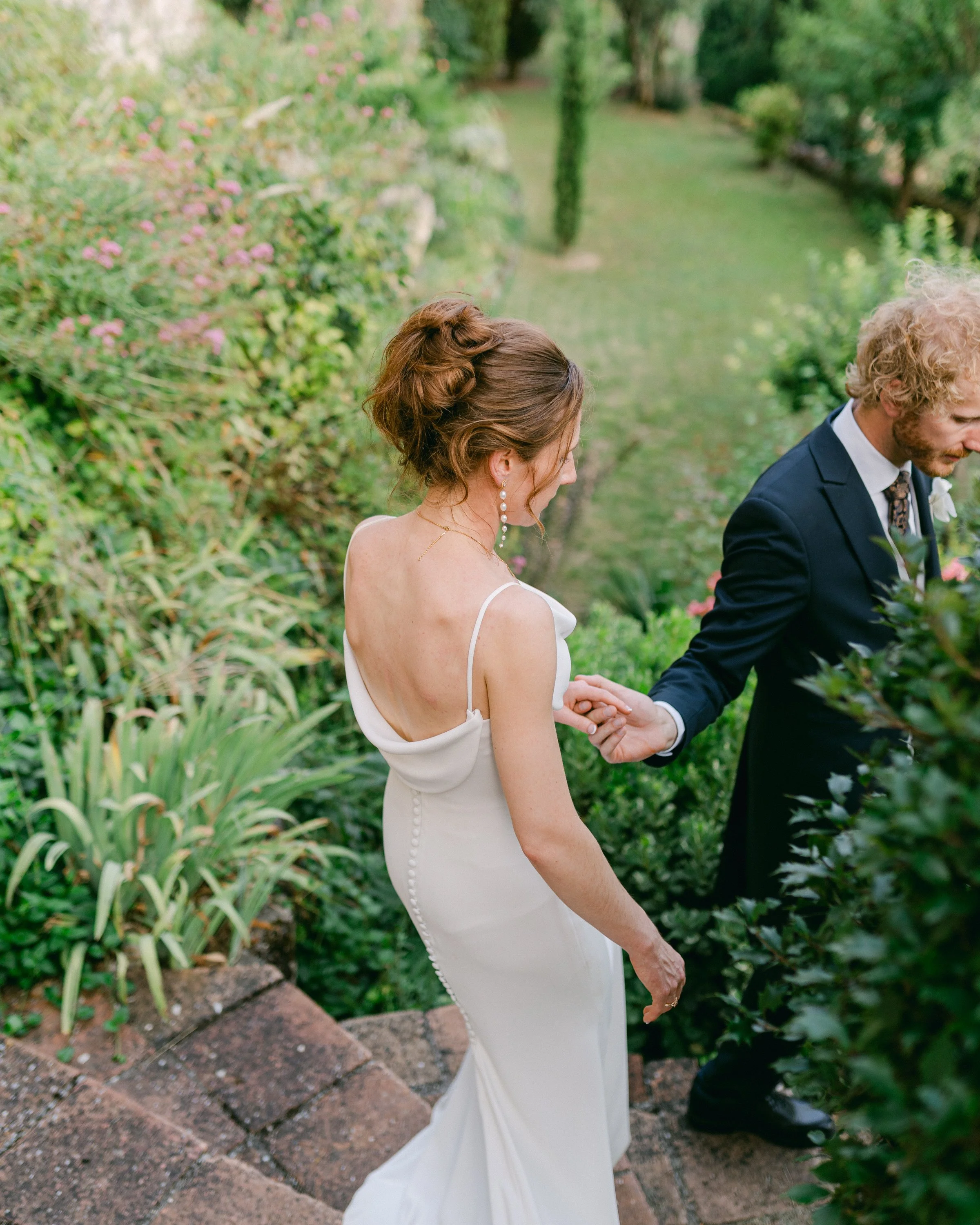 Un couple en costume élégants se tenant la main dans un jardin verdoyant, la femme portant une robe blanche et des boucles d'oreilles pendantes, le homme portant un costume noir.