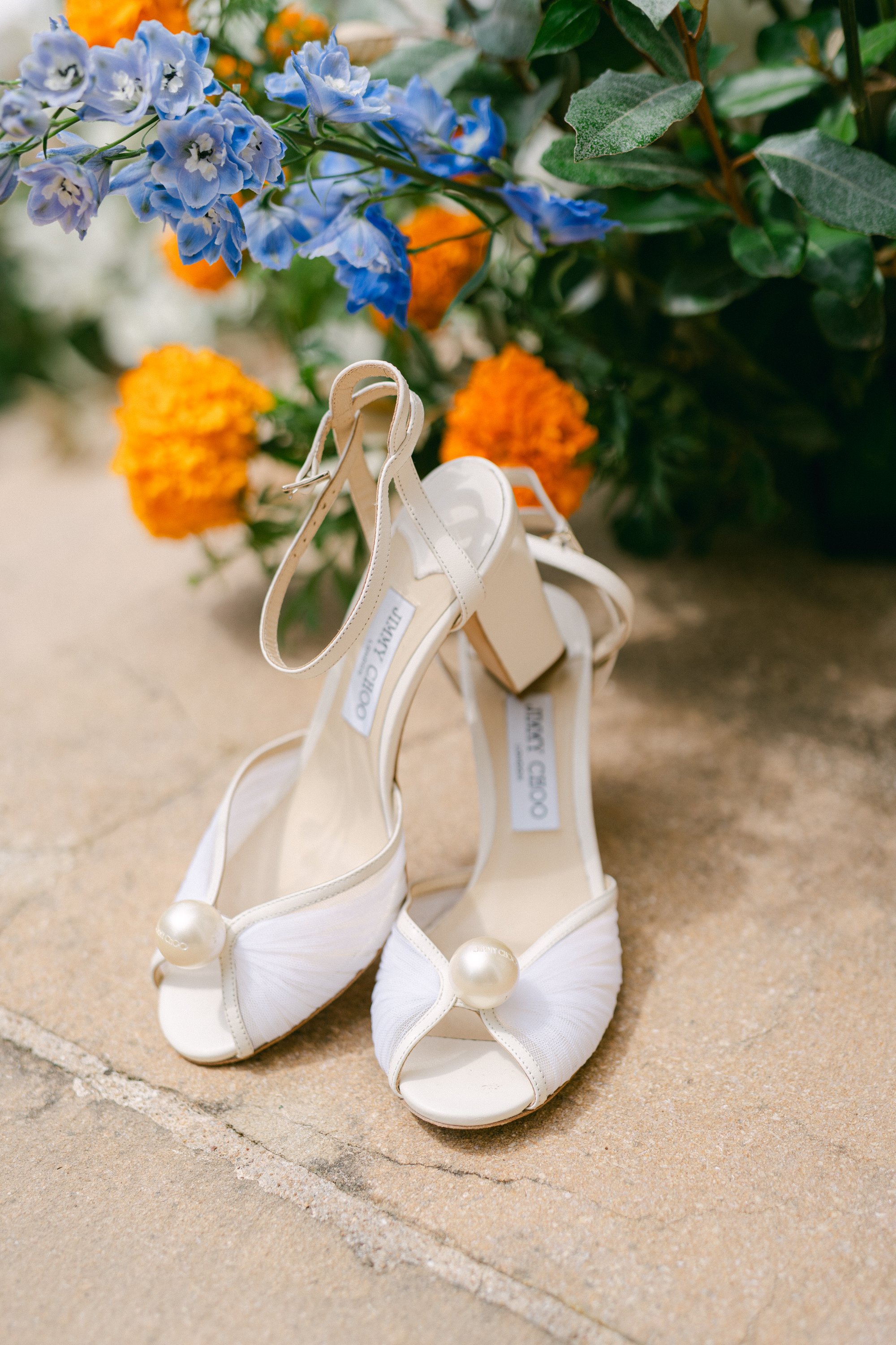 Chaussures à talons blancs avec perles, devant un bouquet de fleurs jaunes et bleues.