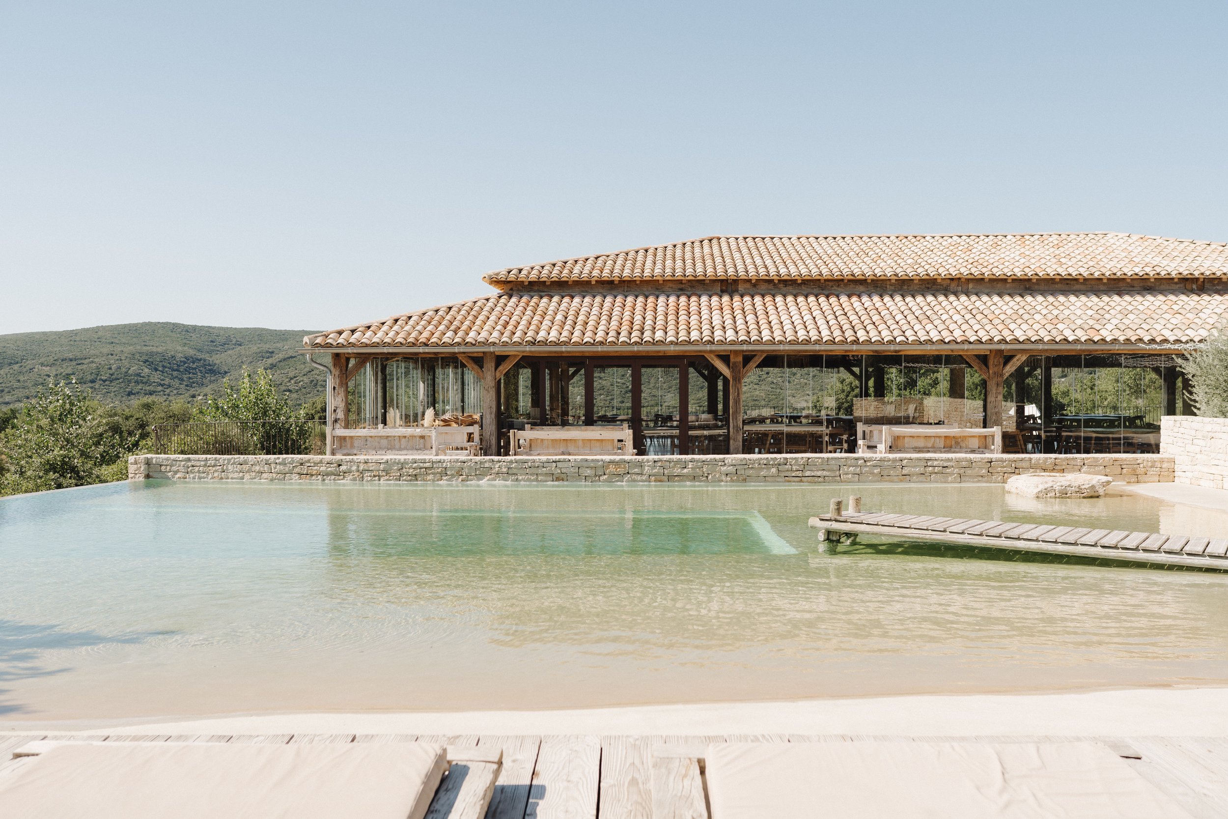 Une piscine à débordement devant un bâtiment en pierre avec un toit en tuiles, entourée de paysages de montagnes et de verdure.