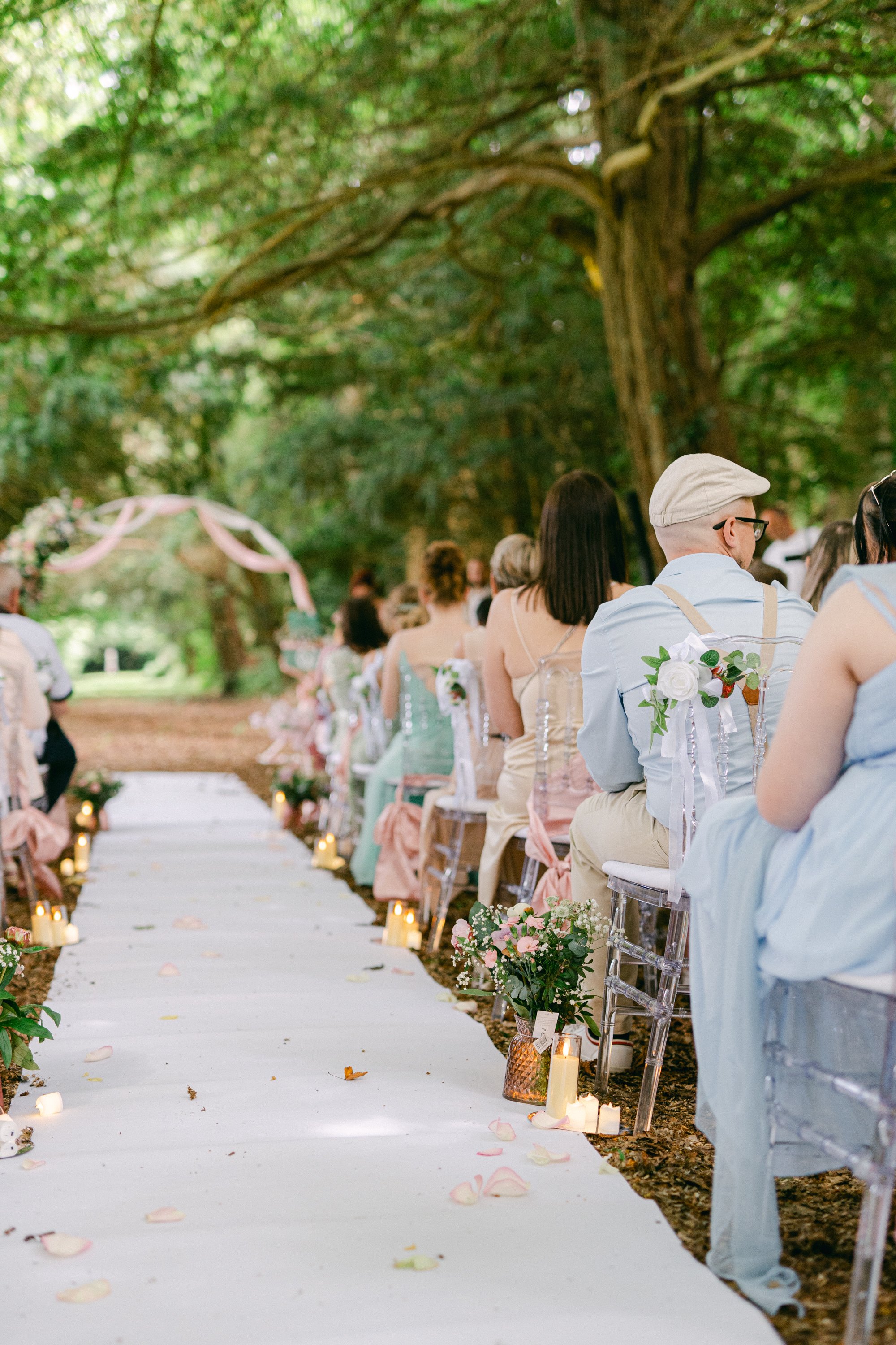 Un mariage en plein air dans une forêt, avec des invités assis sur des chaises en cristal, une allée ornée de fleurs et de bougies, des décorations florales et un arc de mariage décoré de rubans et de fleurs en arrière-plan.