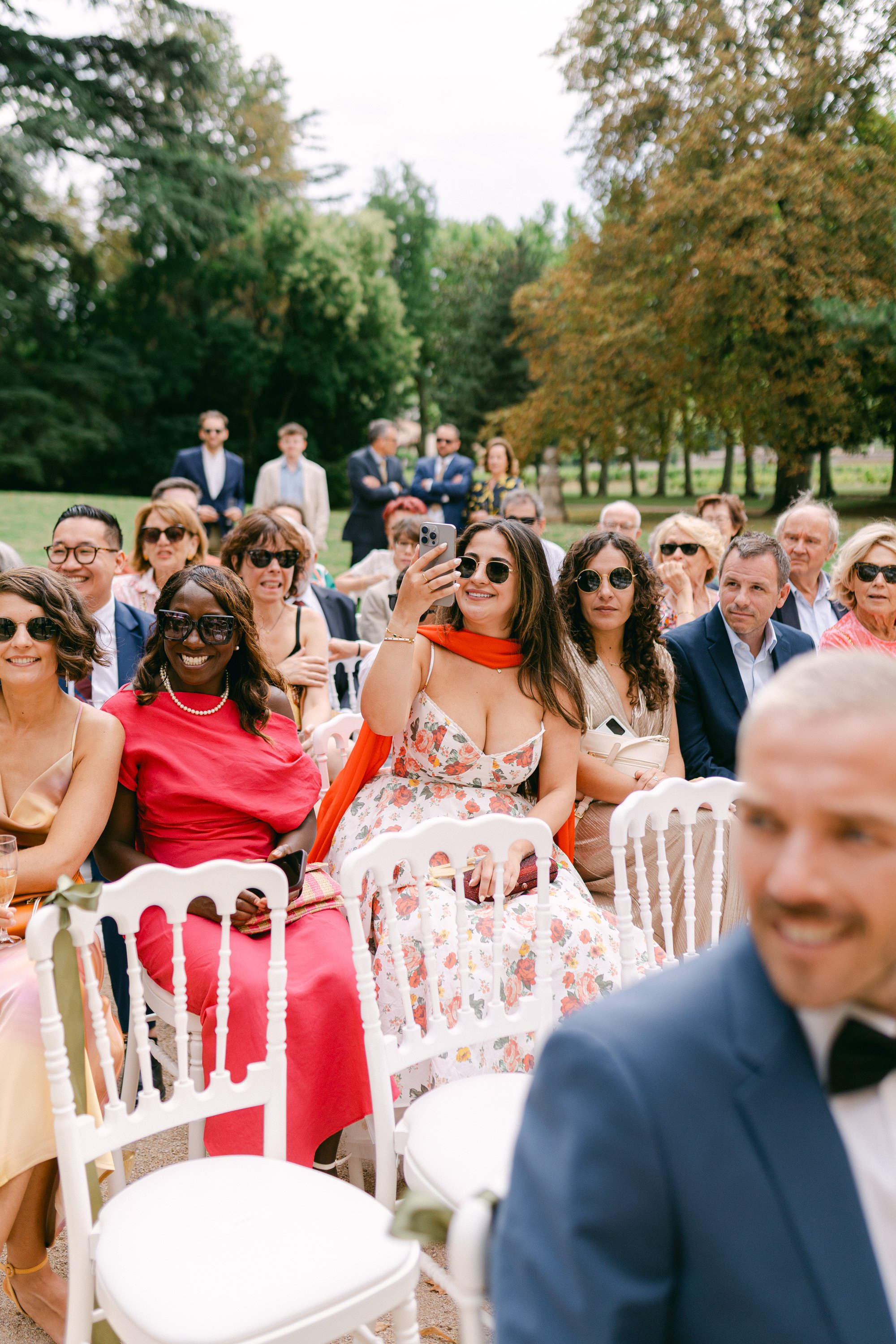 Groupe de personnes assises à un événement en plein air, quelques-unes portent des lunettes de soleil, et une femme en milieu de photo prend une photo avec son téléphone.