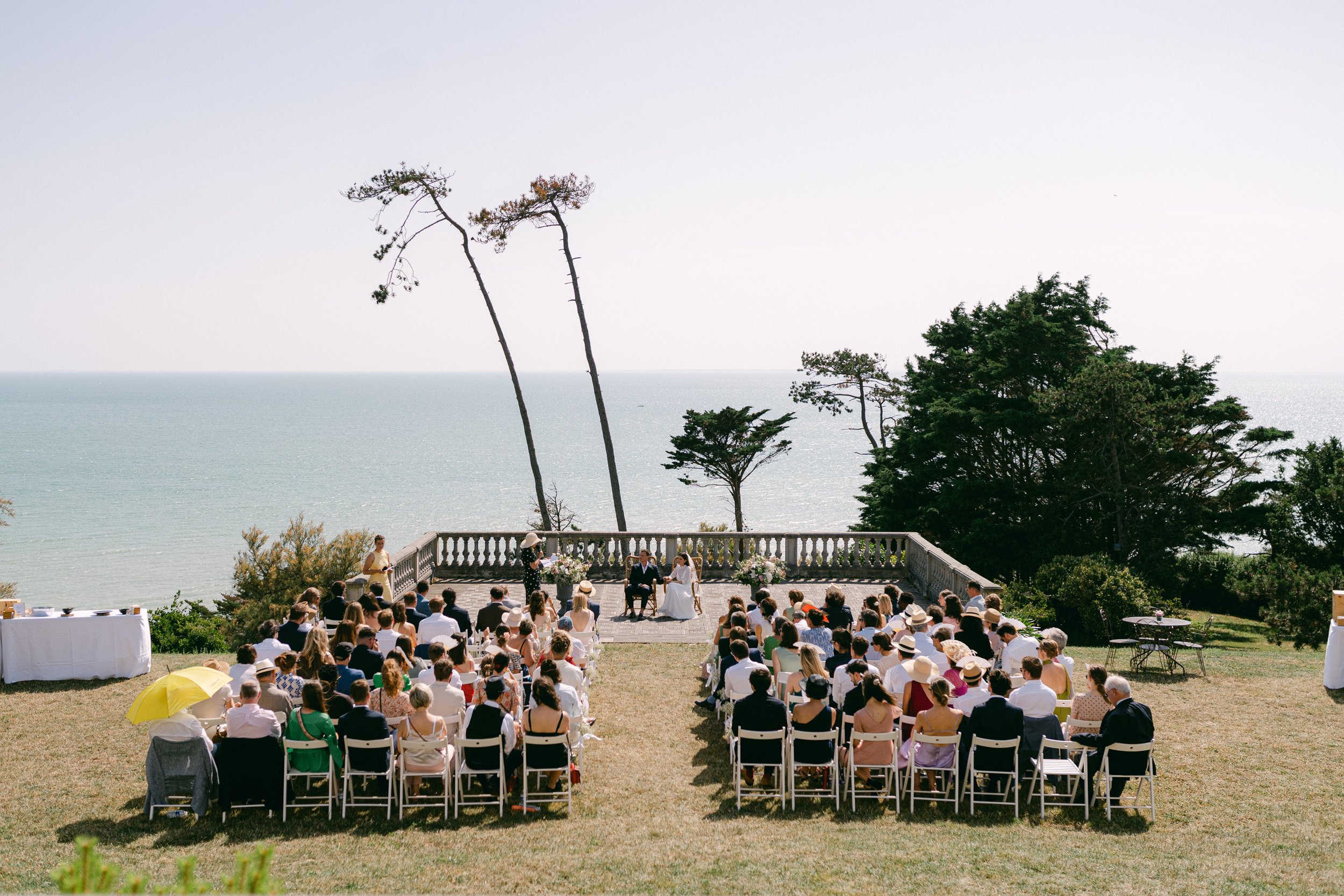 Cérémonie de mariage en plein air face à la mer, avec des invités assis sur des chaises blanches, un couple de mariés devant un officiant, et un décor naturel avec des arbres et un ciel ensoleillé.