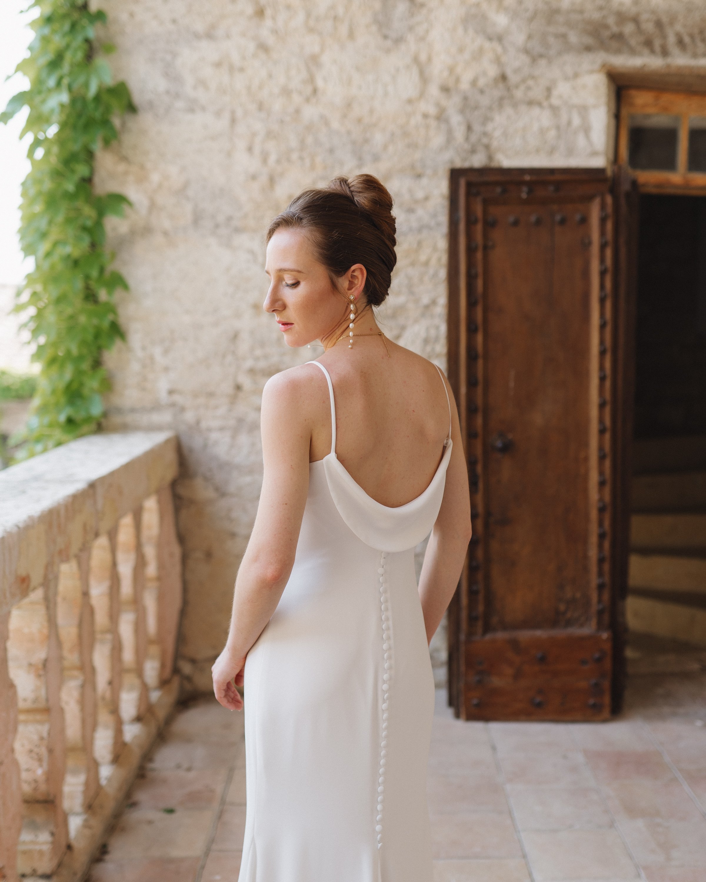 Une femme porte une robe de mariage blanche avec un décolleté drapé dans un lieu en pierre avec des volets en bois.
