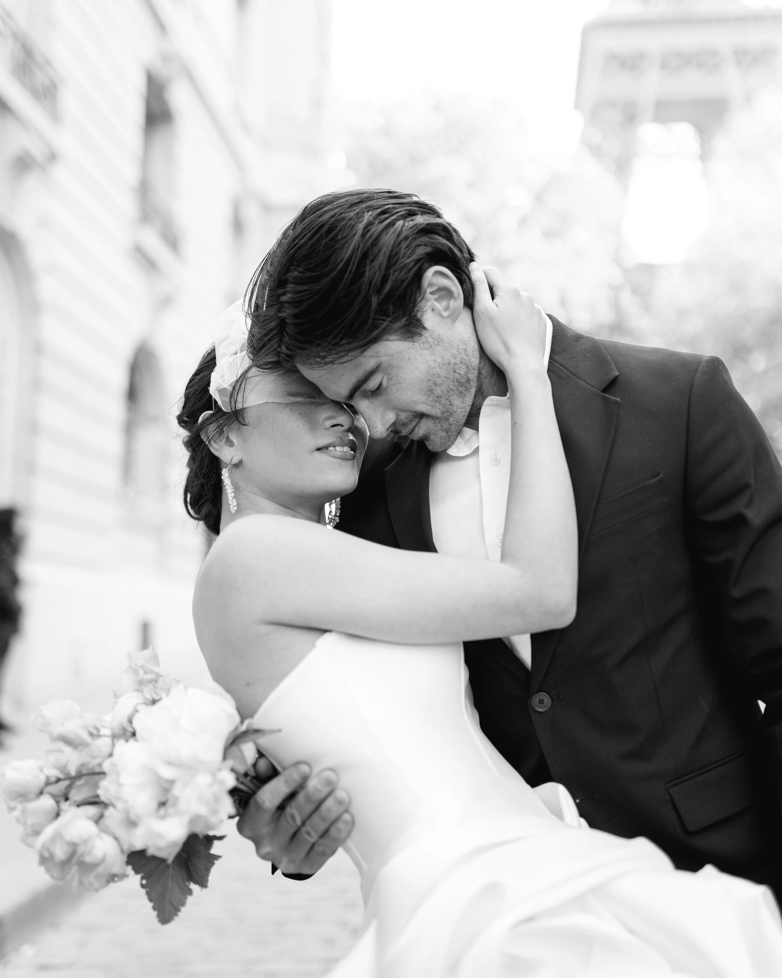 Un couple en mariage, en noir et blanc, partage un moment intime, la femme vêtue d'une robe de mariée et le homme en costume, dans une ambiance romantique en extérieur.