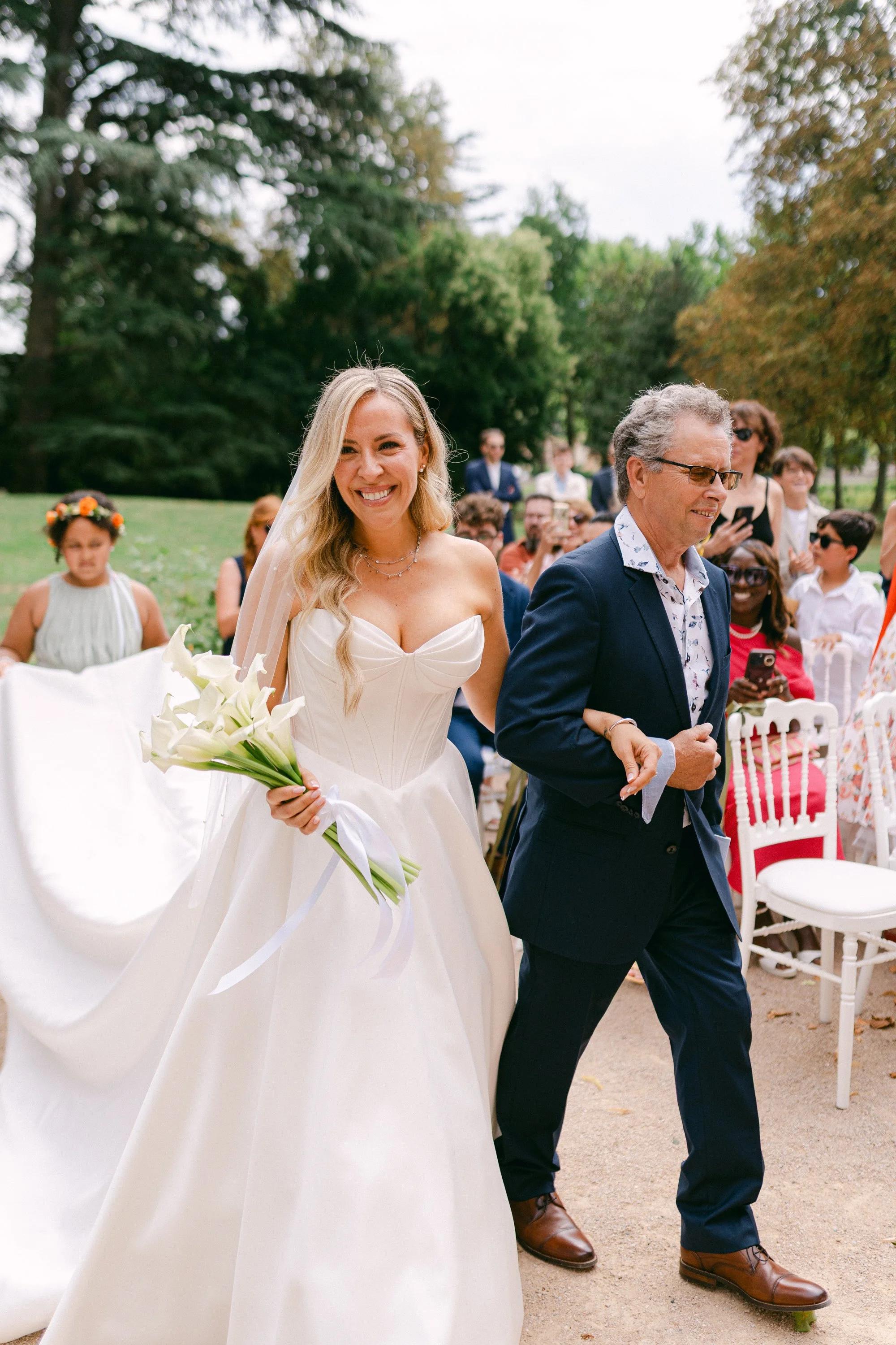 Une mariée souriante en robe blanche, tenant un bouquet de calla lily, marche dans un jardin lors d'un mariage, accompagnée d'un homme en costume.