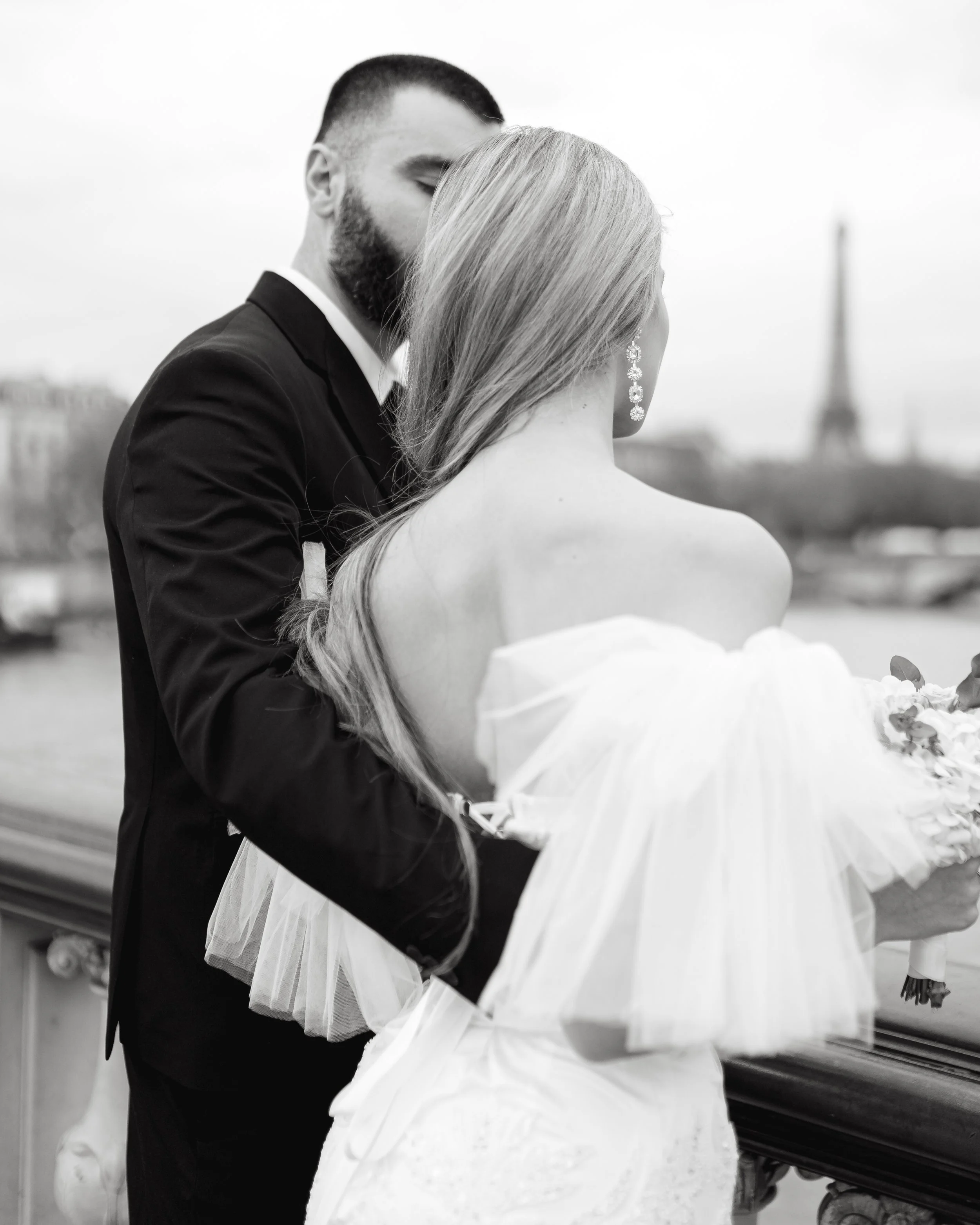 Un couple de mariage posant sur un pont avec la Tour Eiffel en arrière-plan, en noir et blanc.