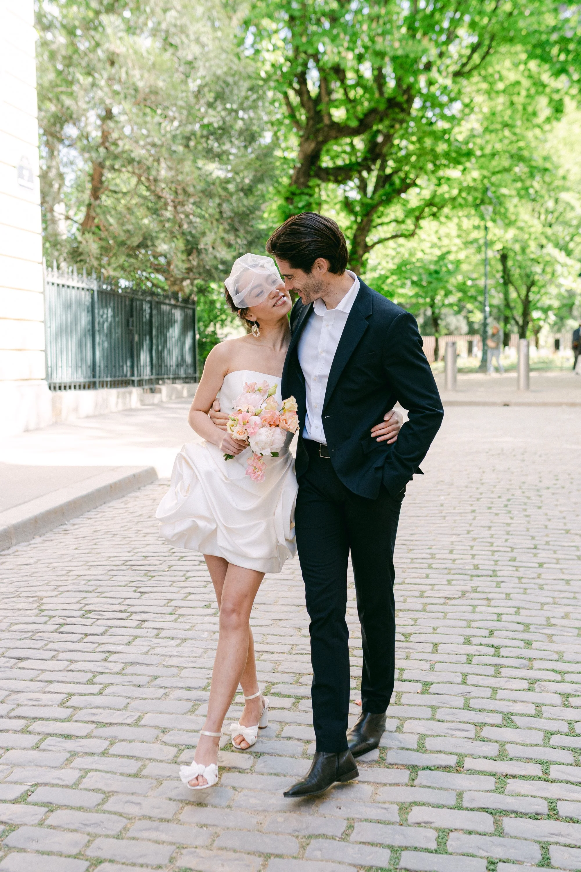 Un couple marié marche ensemble sur une rue pavée, entouré d'arbres verts. La femme porte une robe de mariage blanche, un voile léger, des chaussures à talons et tient un bouquet de fleurs roses. L'homme porte un costume noir, une chemise blanche et des bottines noires.