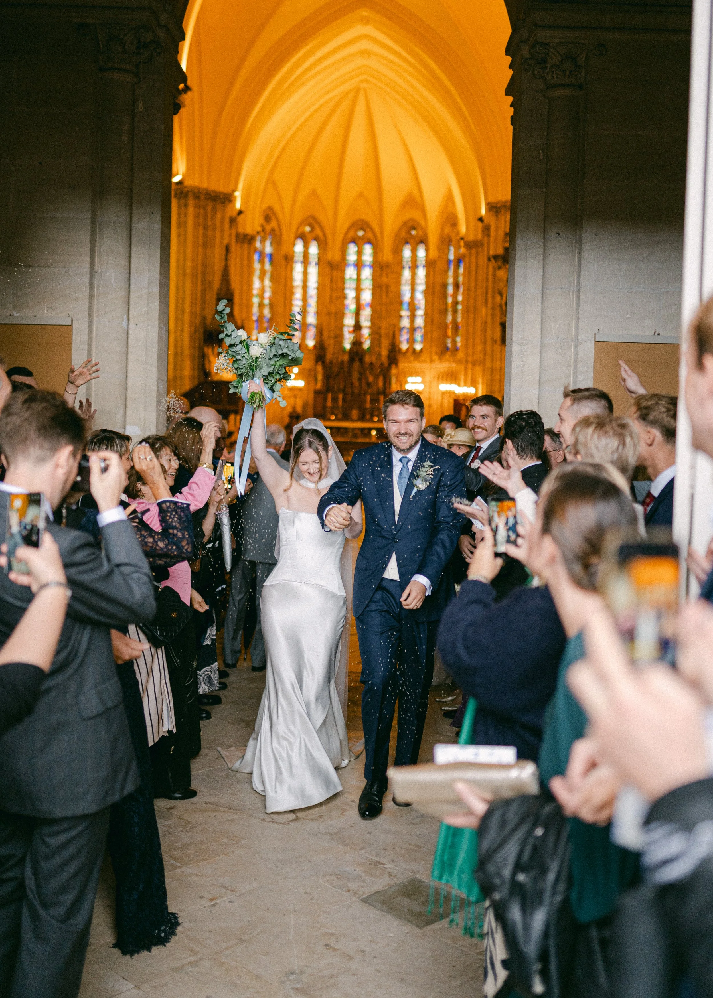 Un couple de mariés entre dans une église après leur mariage, entourés de proches applaudissant et prenant des photos. La mariée porte une robe blanche et le marié un costume bleu.