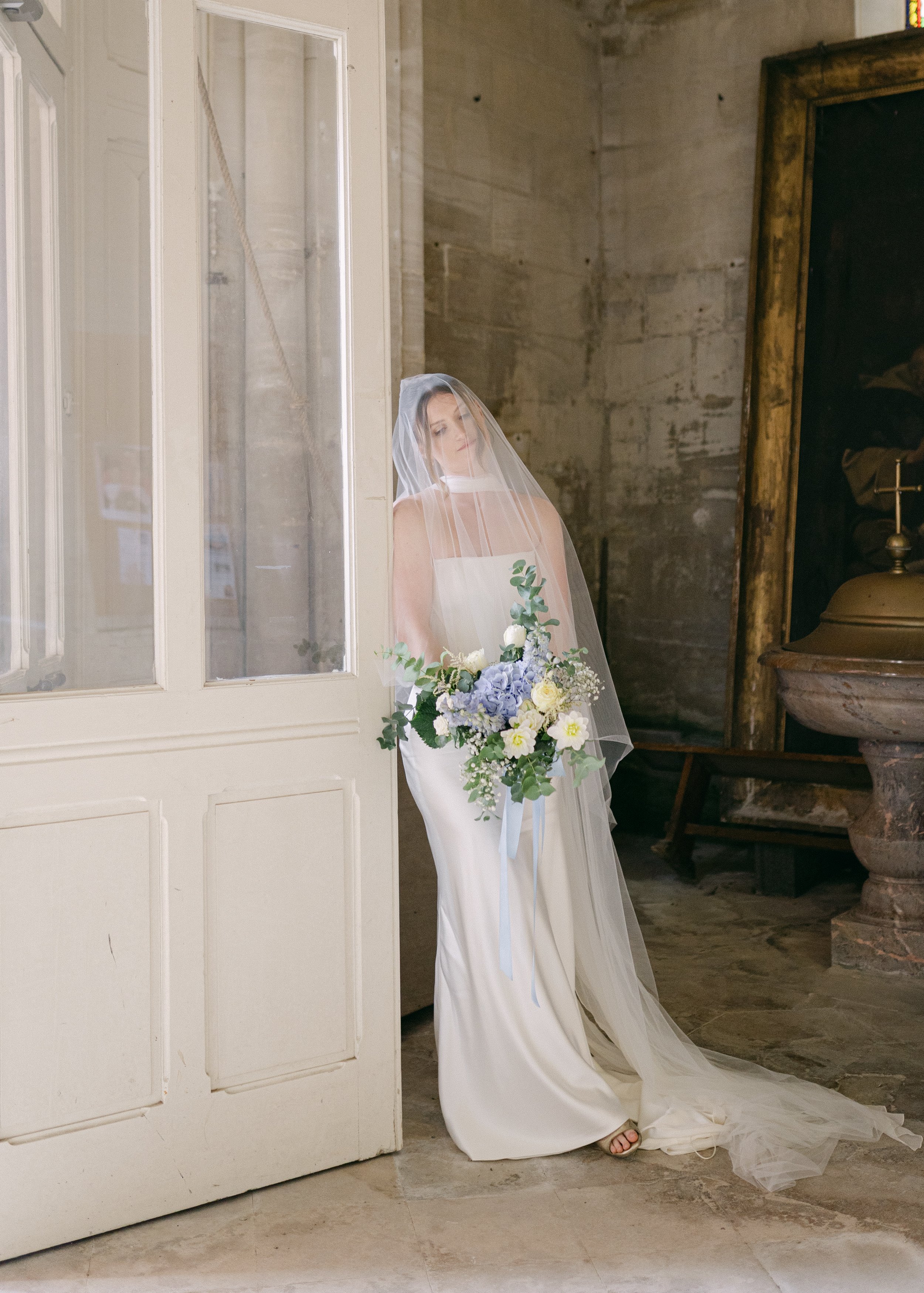 Une mariée en robe blanche portant un voile, tenant un bouquet de fleurs blanches, violettes et vertes dans un lieu au décor ancien en pierre, avec une porte en bois peint en blanc.