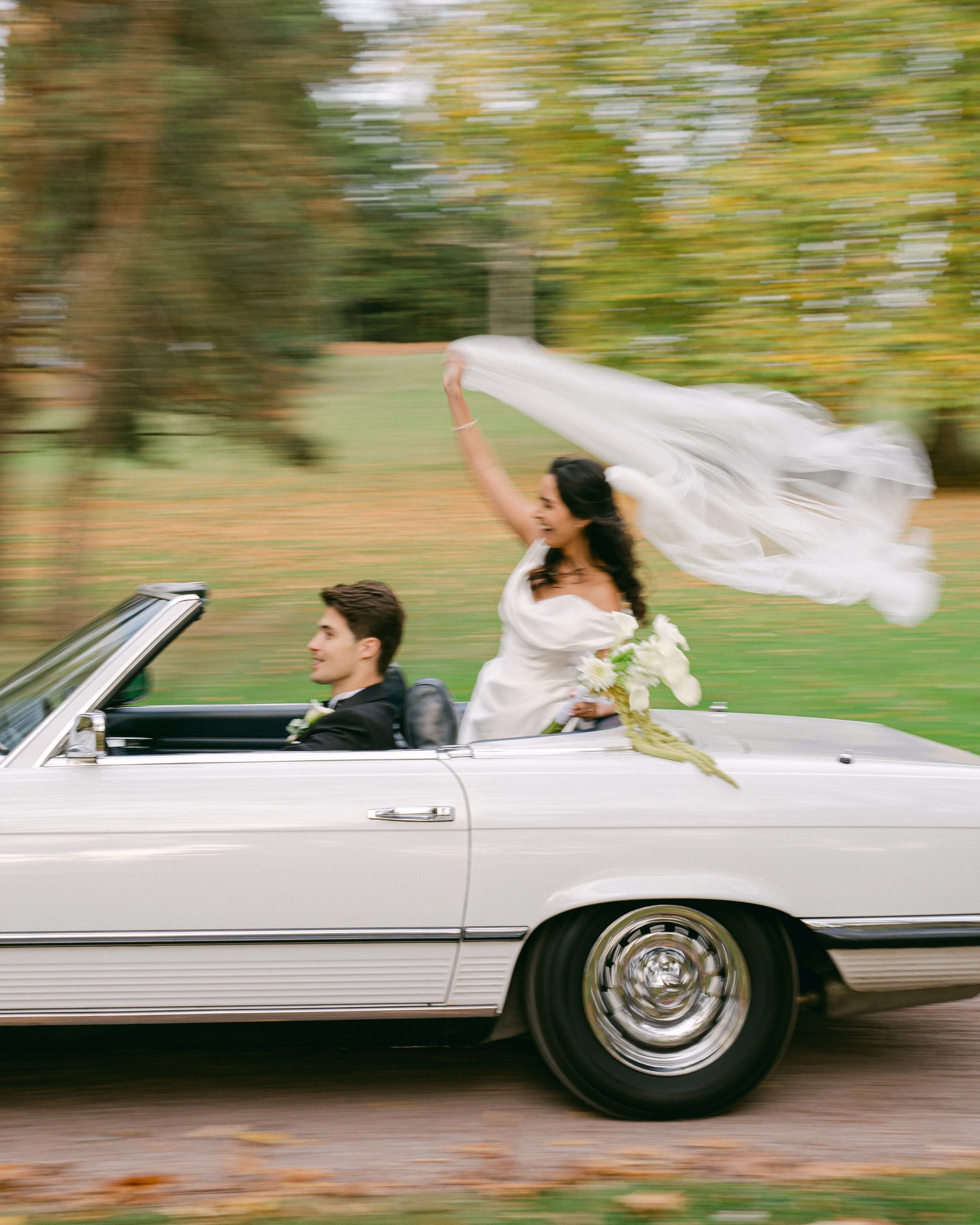 Une paire mariée dans une voiture décapotable blanche en marche, la femme brandissant une voilette et tenant un bouquet de fleurs