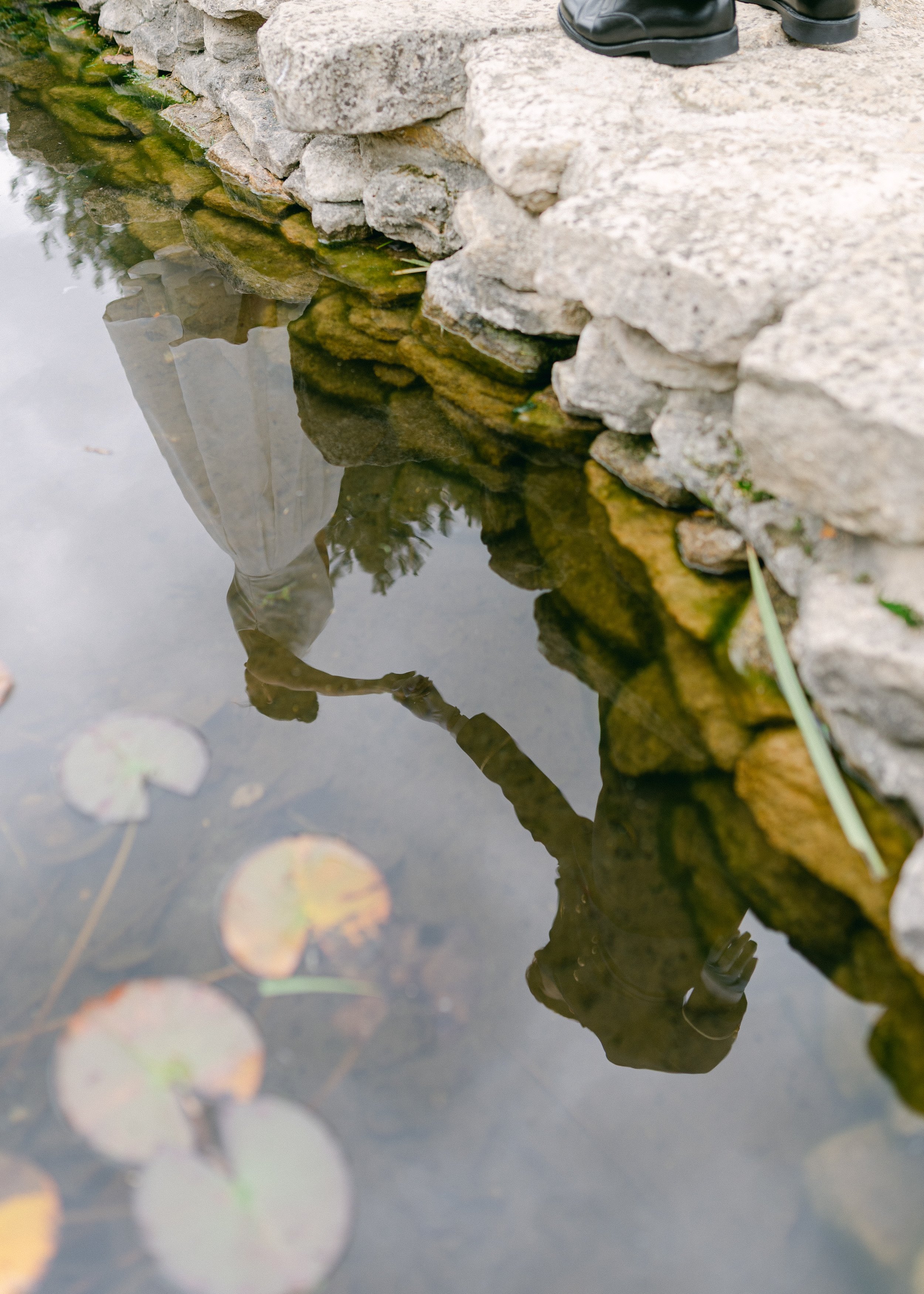 Reflet d'une personne portant un manteau gris et un pantalon gris dans l'eau d'un étang, avec des nénuphars flottants, au bord d'un petit pont en pierre.