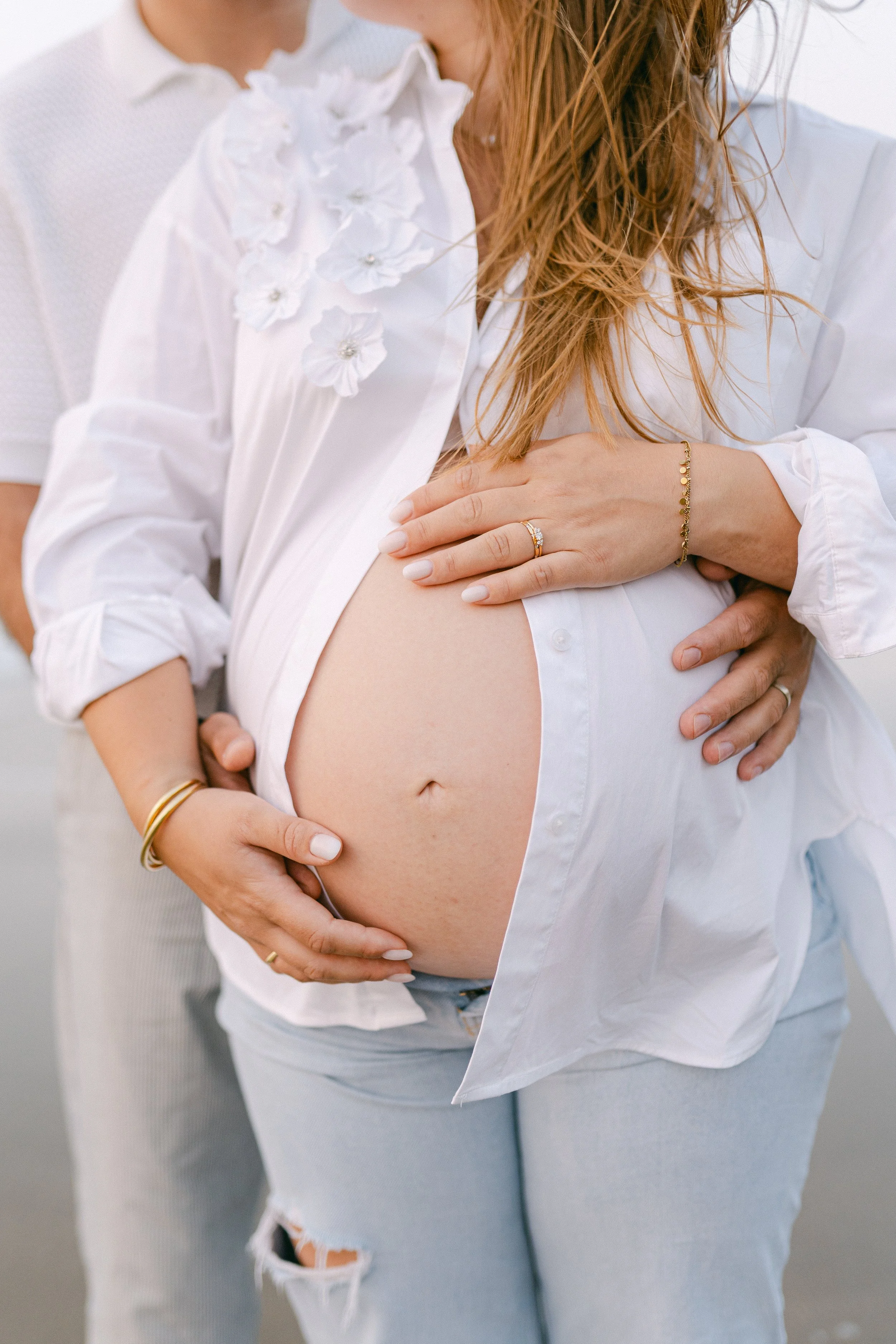 Une femme enceinte avec un haut blanc entrouvert et un homme derrière elle, tenant son ventre. La femme porte des bijoux dorés et une chemise blanche décorée de fleurs.