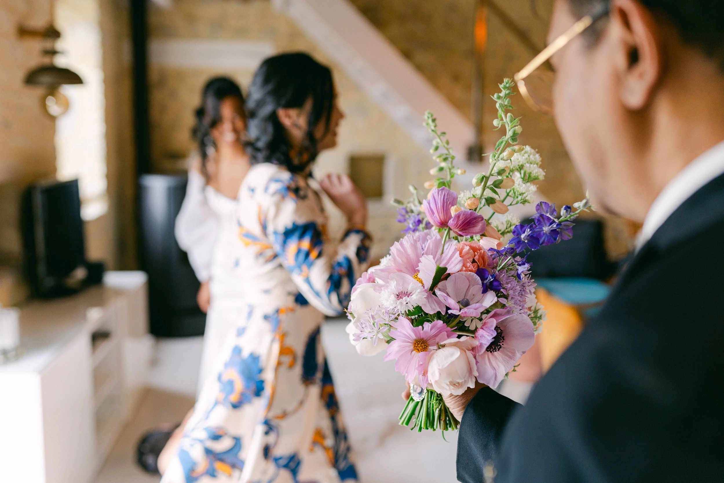 Une personne en costume noir tient un bouquet de fleurs colorées, avec deux autres femmes floues en arrière-plan dans un intérieur.