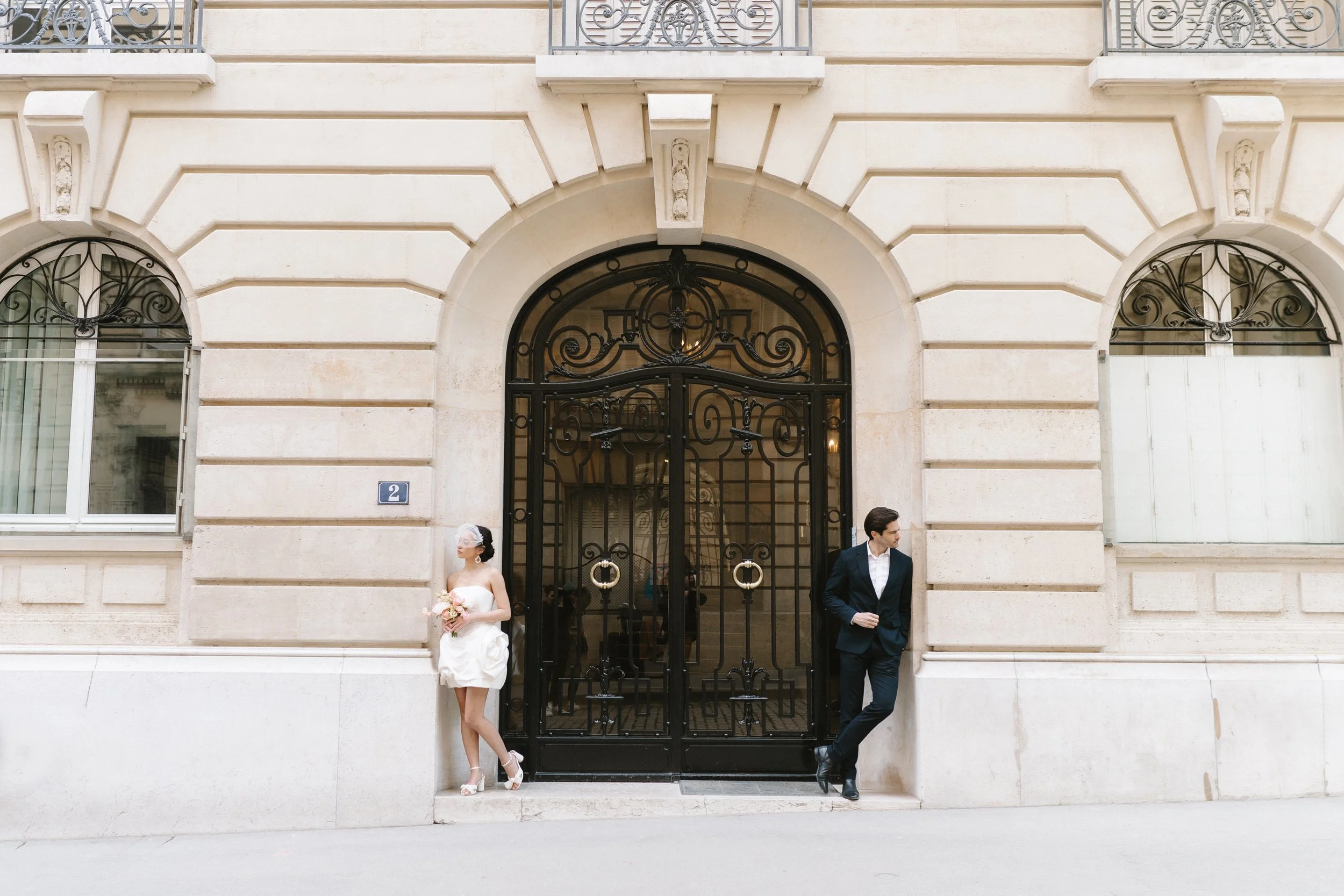 Un couple en vêtements de mariage se tient de chaque côté d’un portail en fer noir, dans une rue urbaine. La femme porte une robe de mariée blanche et trace une ligne avec ses chaussures à talons hauts, tandis que l’homme porte un costume sombre et a