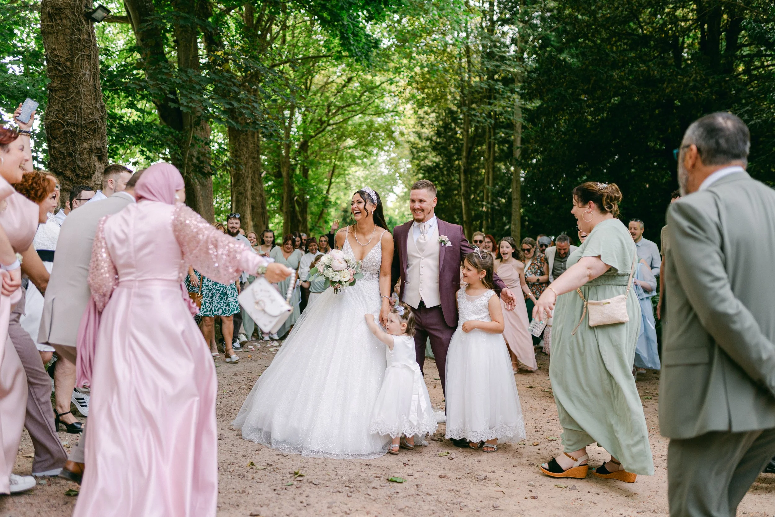 Mariage en plein air dans une forêt. Le couple de mariés, la mariée en robe blanche et le marié en costume sombre, marche main dans la main entourés d'invités heureux en tenue variée.