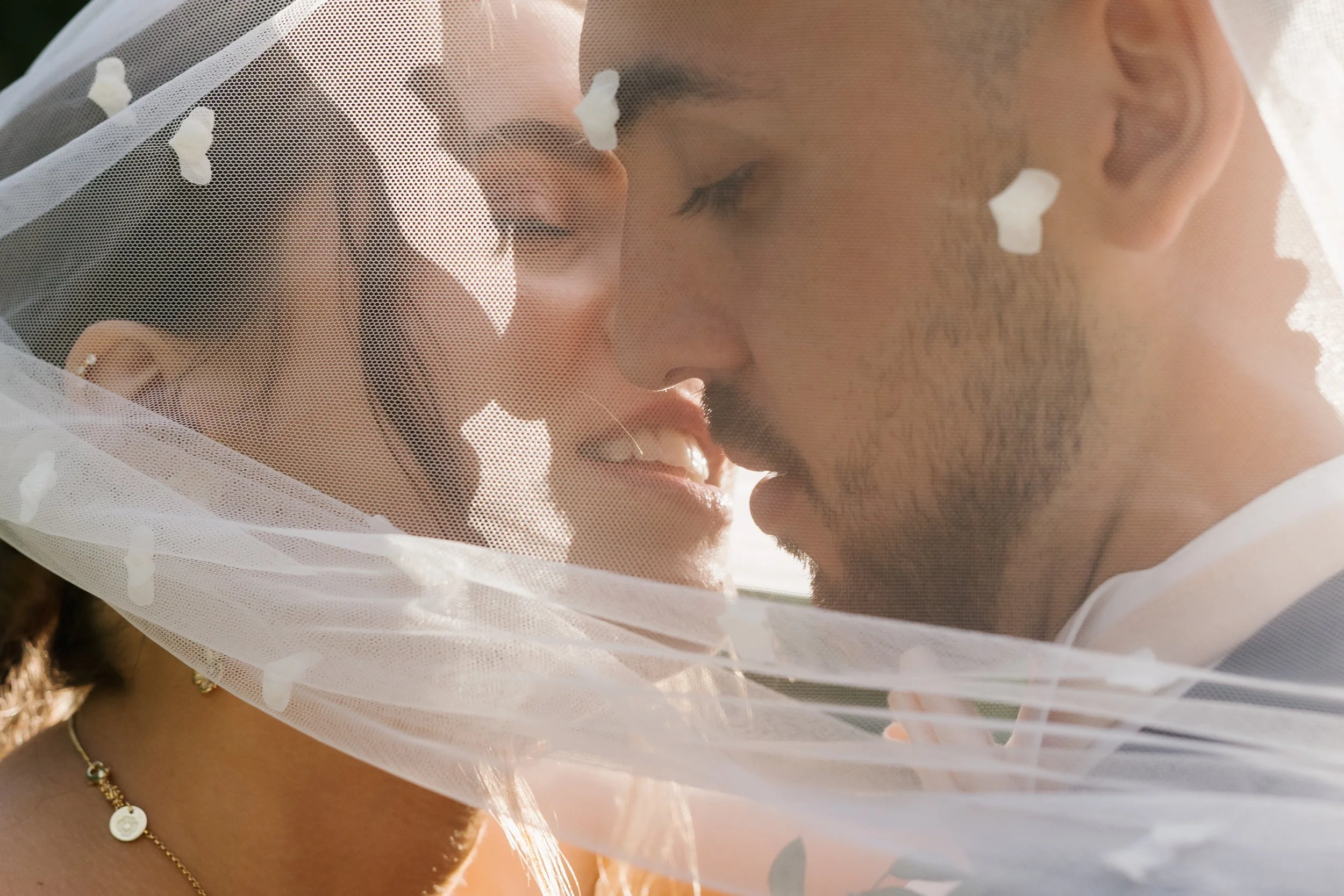 Un couple marié en proximity, leur visage presque touchant, sous un voile blanc de mariage, dans une lumière douce.
