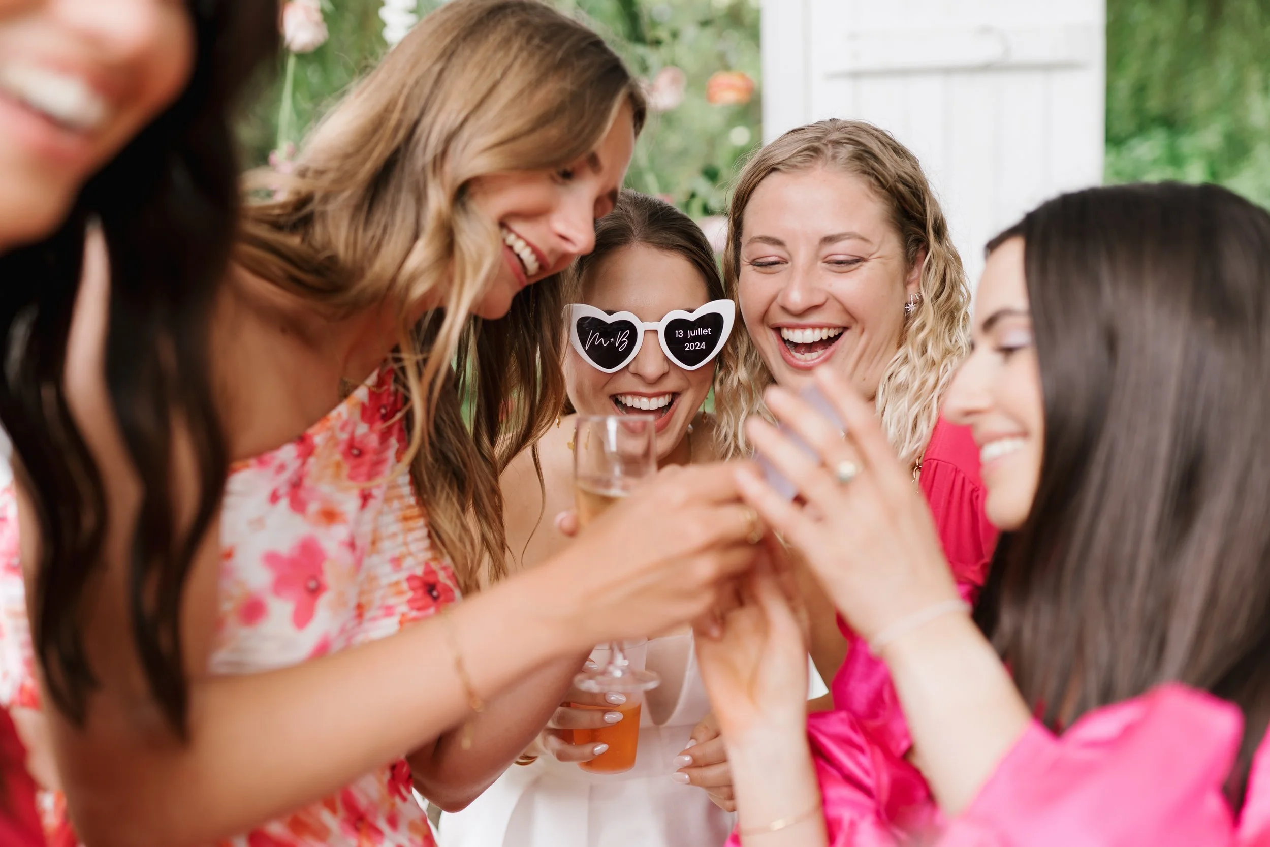 Groupe de femmes souriantes lors d'une célébration, portant des lunettes de soleil en forme de cœur avec la date et un message, tenant un verre avec une boisson à la main, dans un décor extérieur