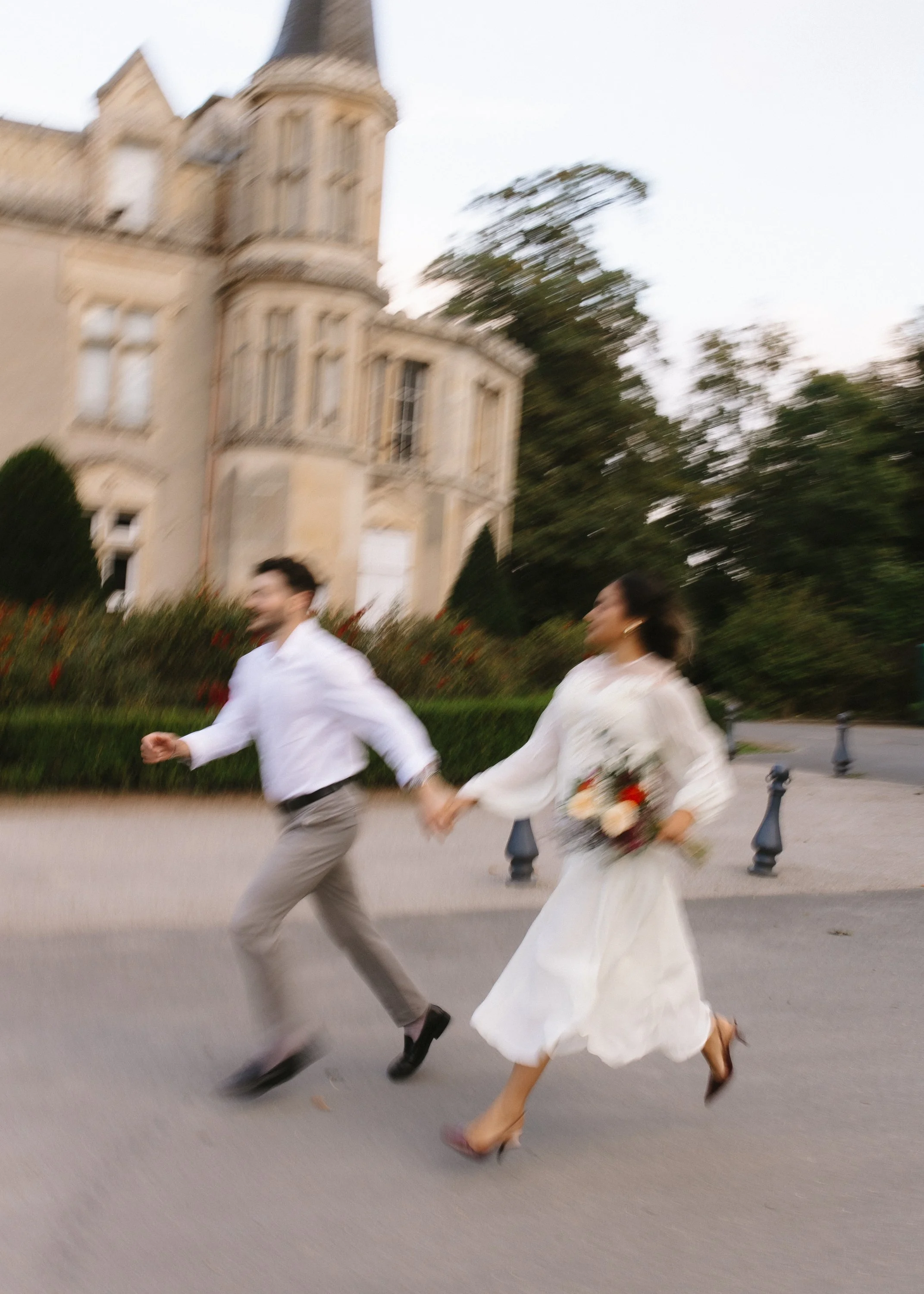 Une femme et un homme, probablement un couple, courant main dans la main devant une grande maison en pierre, la femme tenant un bouquet de fleurs, dans un contexte de style romantique ou lors d'un événement spécial.
