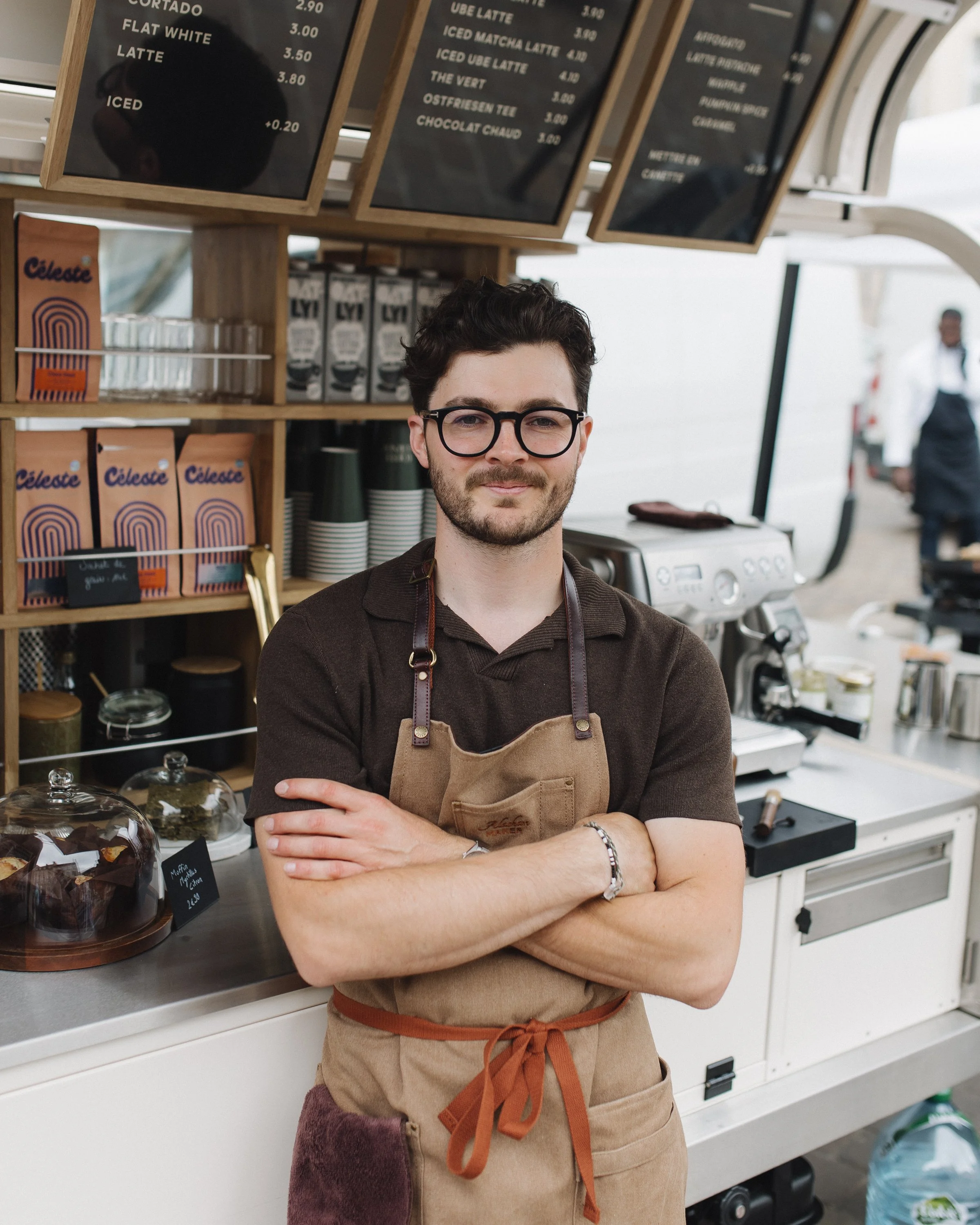Un homme avec des lunettes et une barbe, portant un tablier beige, se tient derrière un comptoir dans un café ou un stand de vente, avec une machine à café et une ardoise au fond.