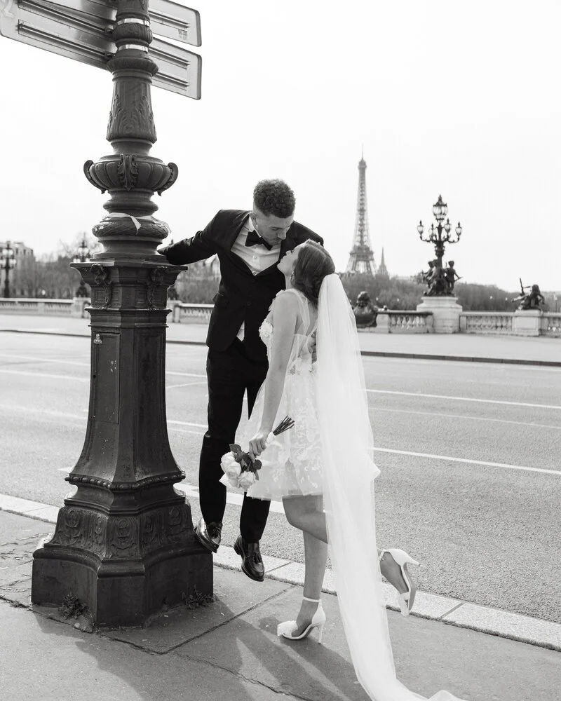 Un couple de mariés en robe de mariée et costume, posant pour une photo romantique à Paris avec la Tour Eiffel en arrière-plan. La mariée tient un bouquet de fleurs, et ils sont près d'un lampadaire ancien.
