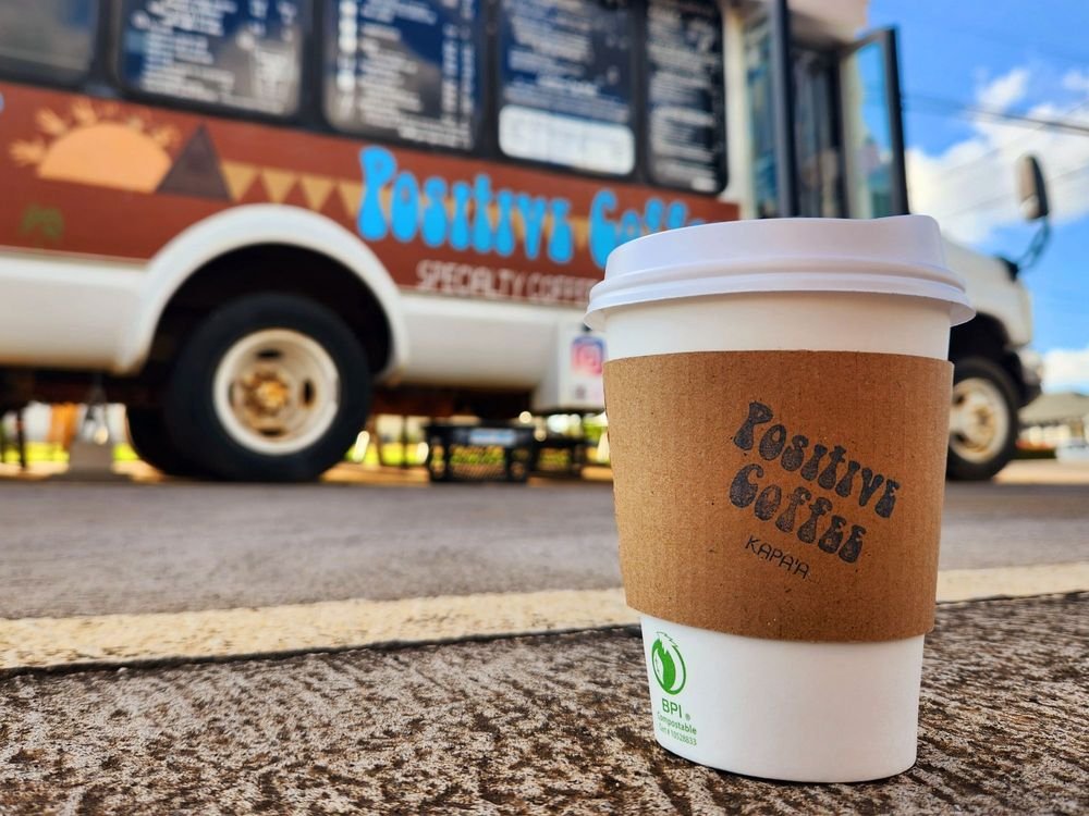 A photo of a paper cup with a Positive Coffee sleeve posed in front of the Positive Coffee bus