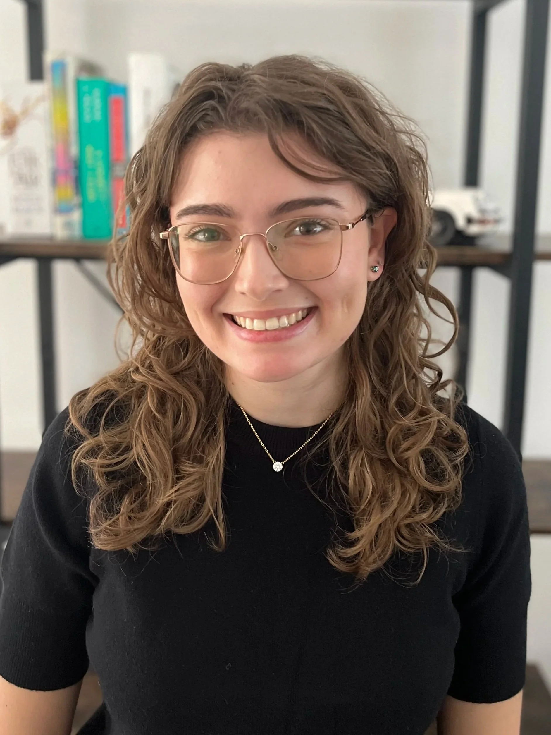 A young woman with curly brown hair and glasses smiling at the camera, wearing a black top and a necklace, sitting in front of a bookshelf.