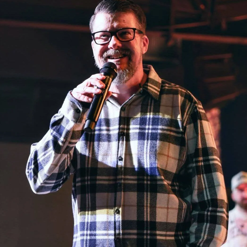 A man with glasses and a beard smiling while holding a microphone, wearing a plaid shirt, in an indoor setting.