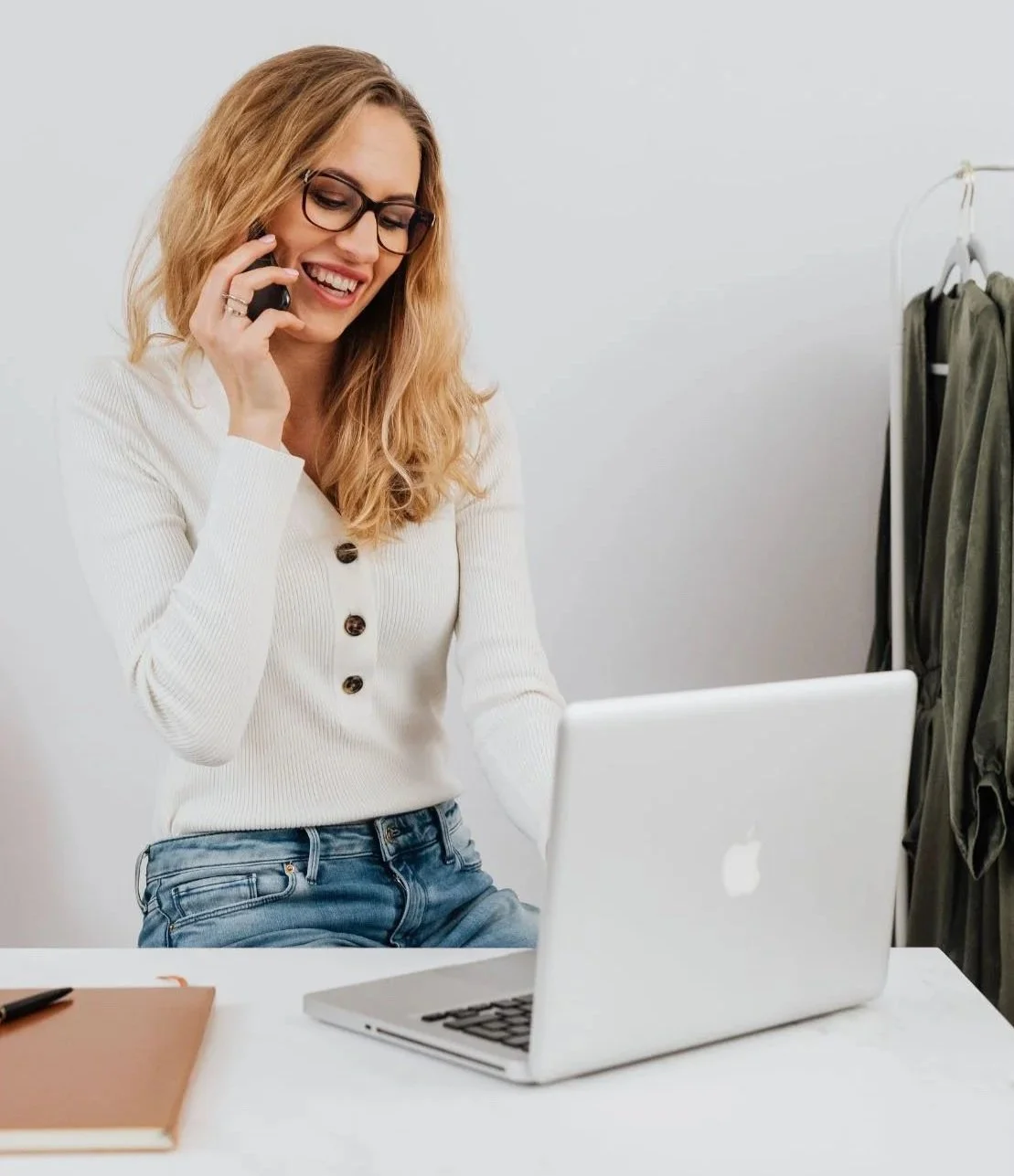 Woman with glasses talking on the phone at a desk with a laptop.