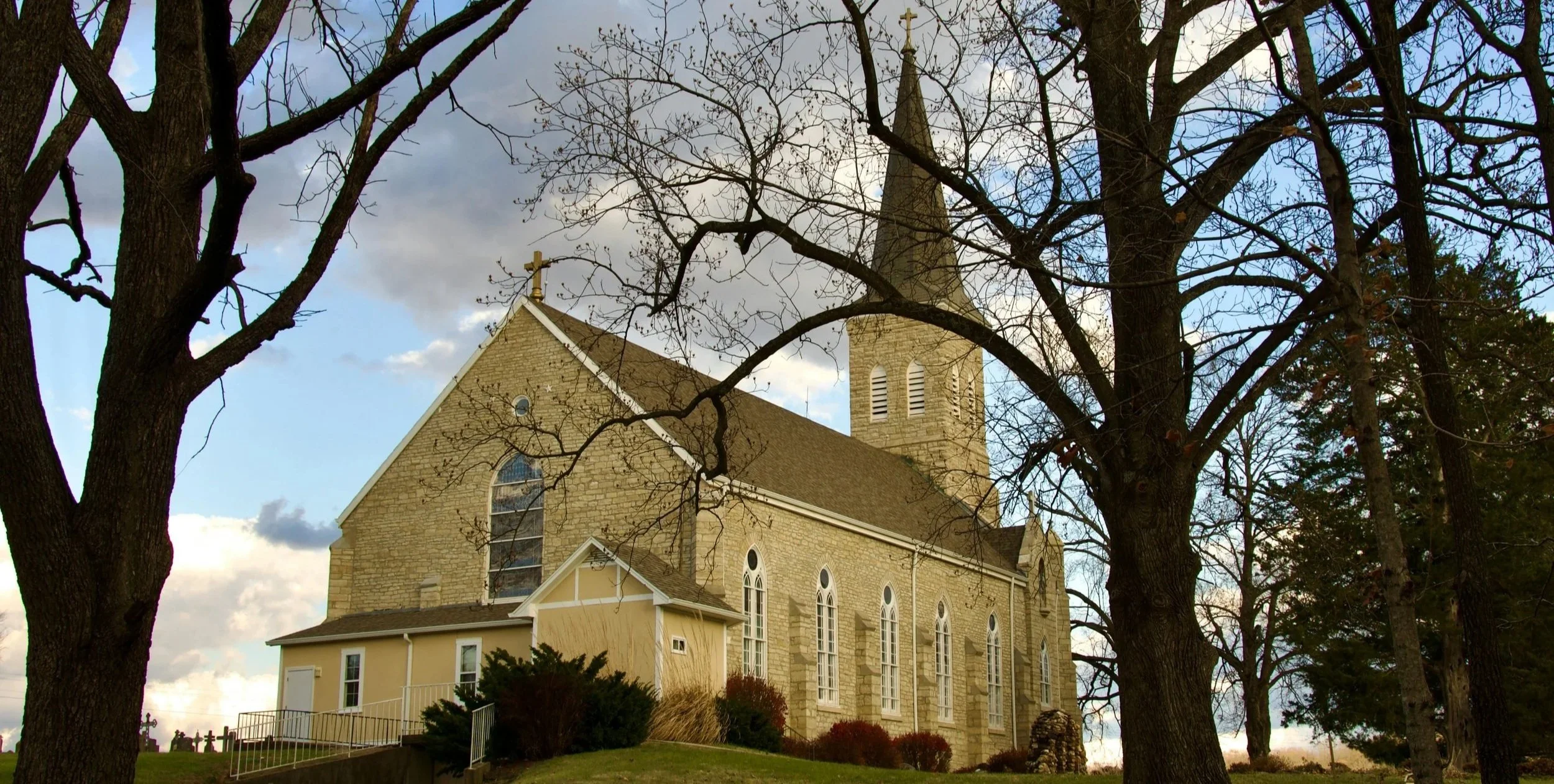 A stone church with a steeple topped by a cross, surrounded by leafless trees, under a cloudy sky.