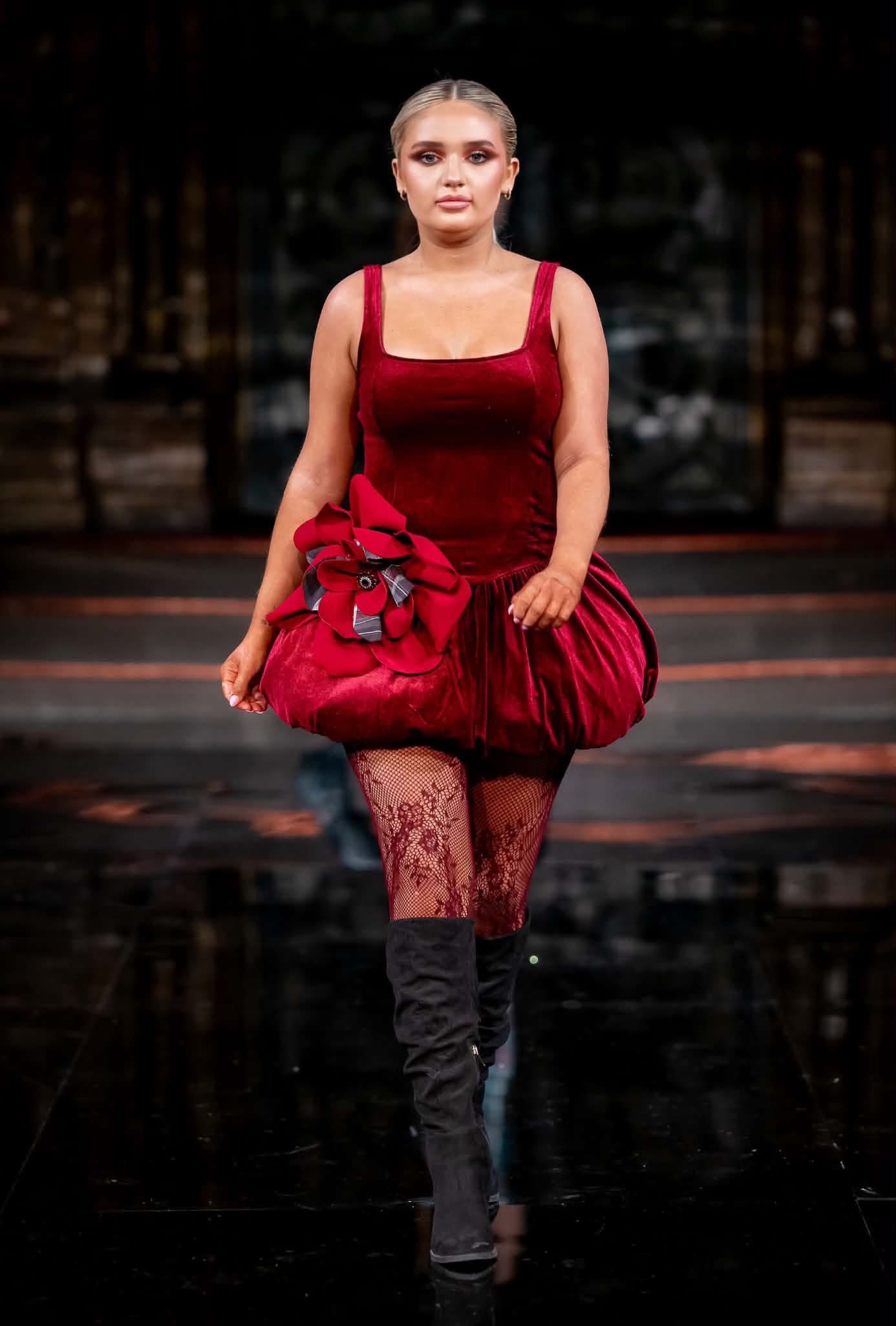 Model walking runway in red dress with floral embellishment, black fishnet stockings, and thigh-high black boots.