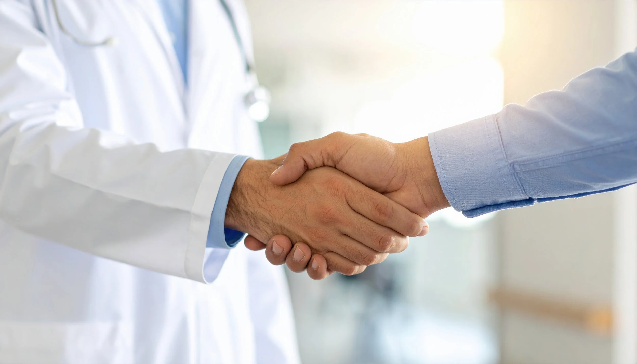 Two people in professional attire shaking hands, one wearing a white lab coat and the other in a light blue shirt, in a well-lit medical or professional setting.