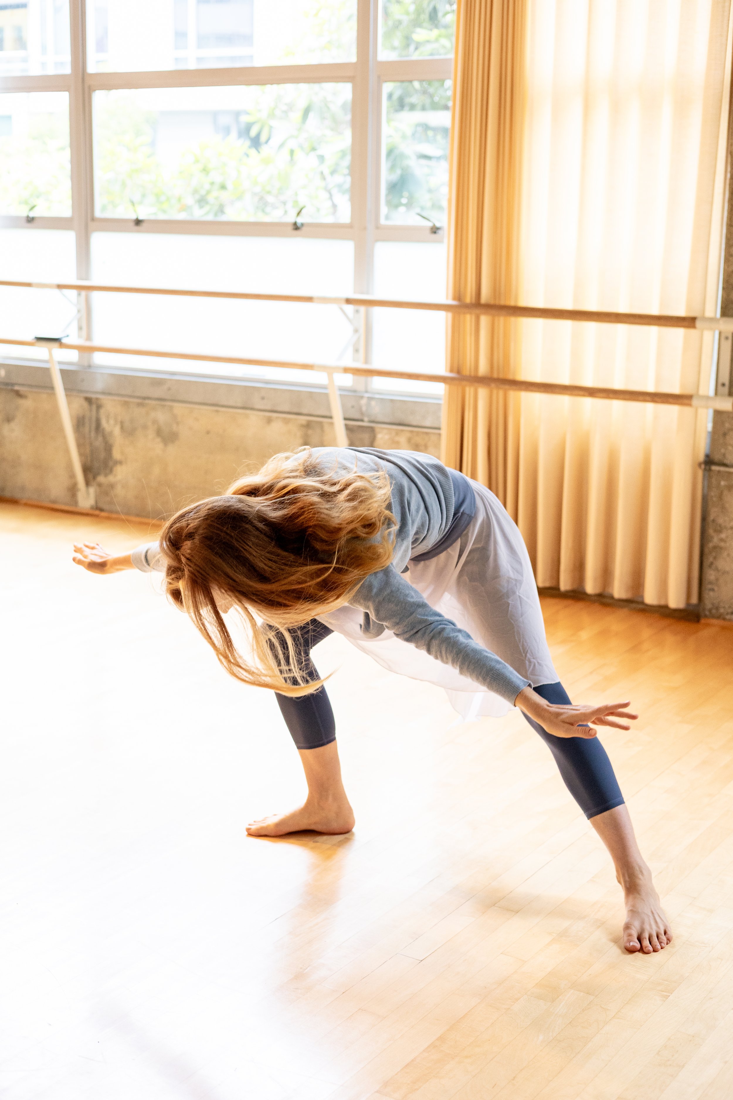 Barefoot dancer bending forward with arms extended in a bright studio with wood floors and large windows